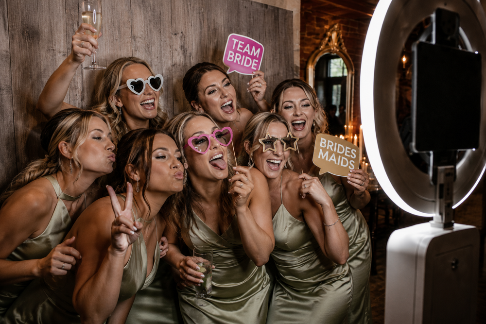 Bridesmaids in sage green dresses pose with fun photo booth props including Team Bride sign and ring light.
