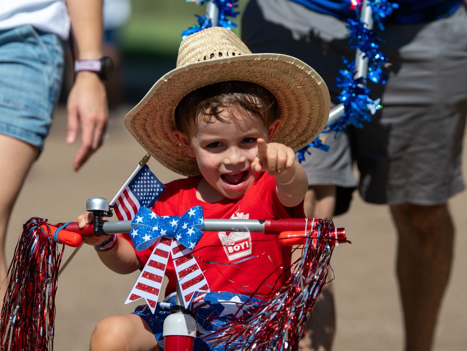 Young child in straw hat rides decorated tricycle with American flags at a 4th of July parade.