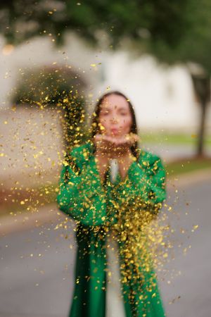 Woman in green dress blowing sparkling gold confetti toward camera outdoors.