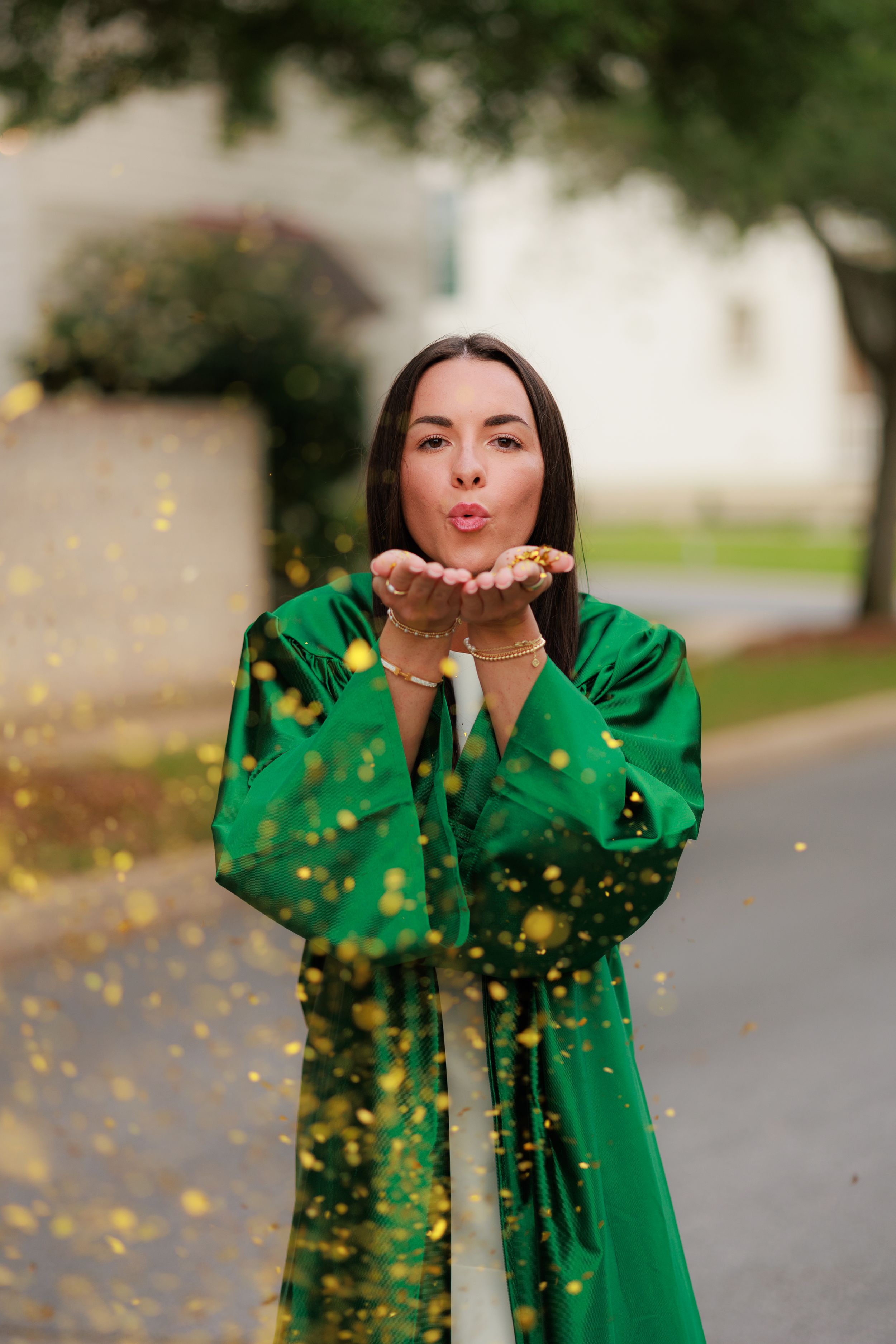 Woman in green graduation gown blowing gold confetti from her hands outdoors.