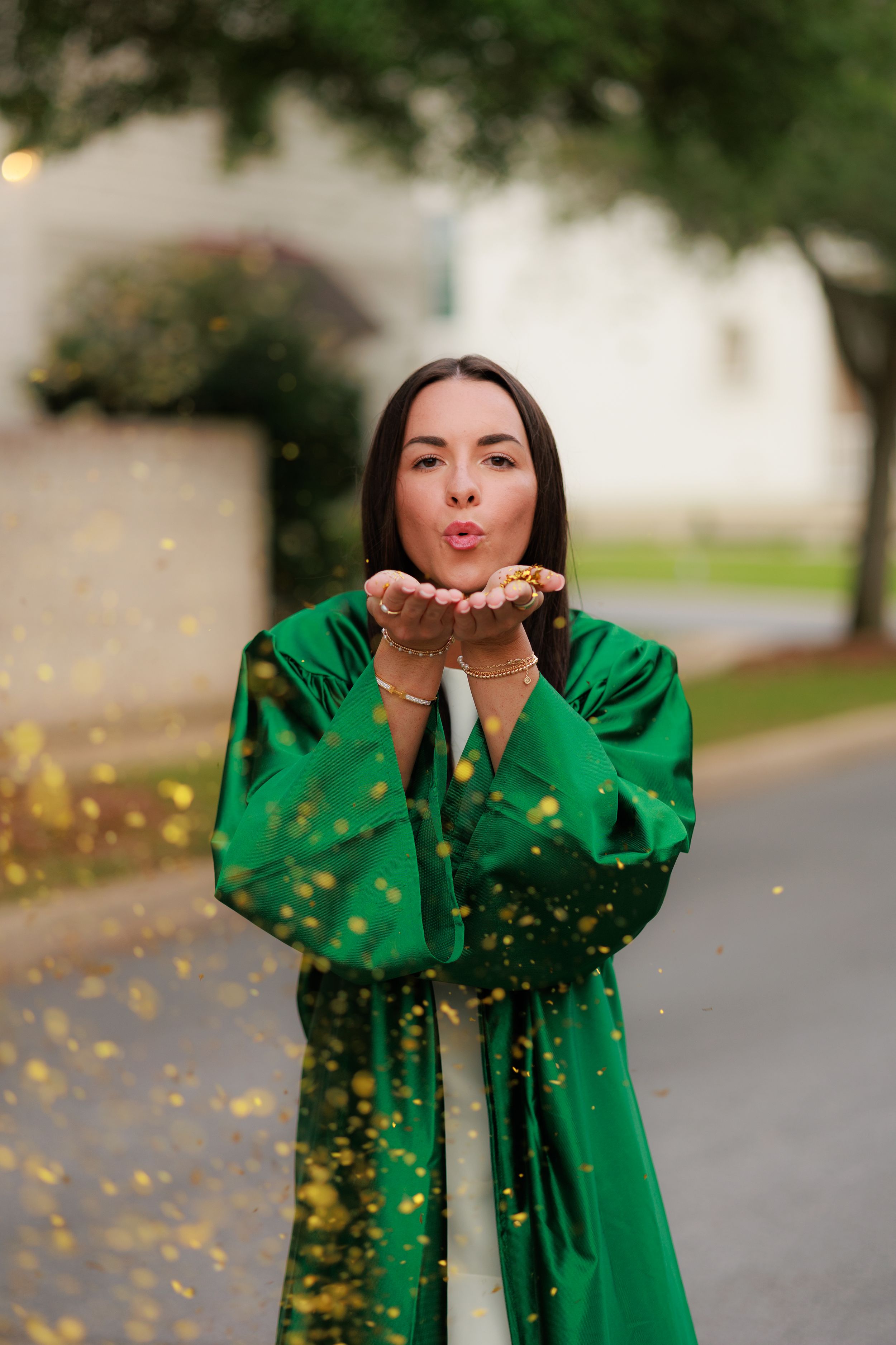 Woman in green graduation gown blowing gold glitter confetti from her hands outdoors.
