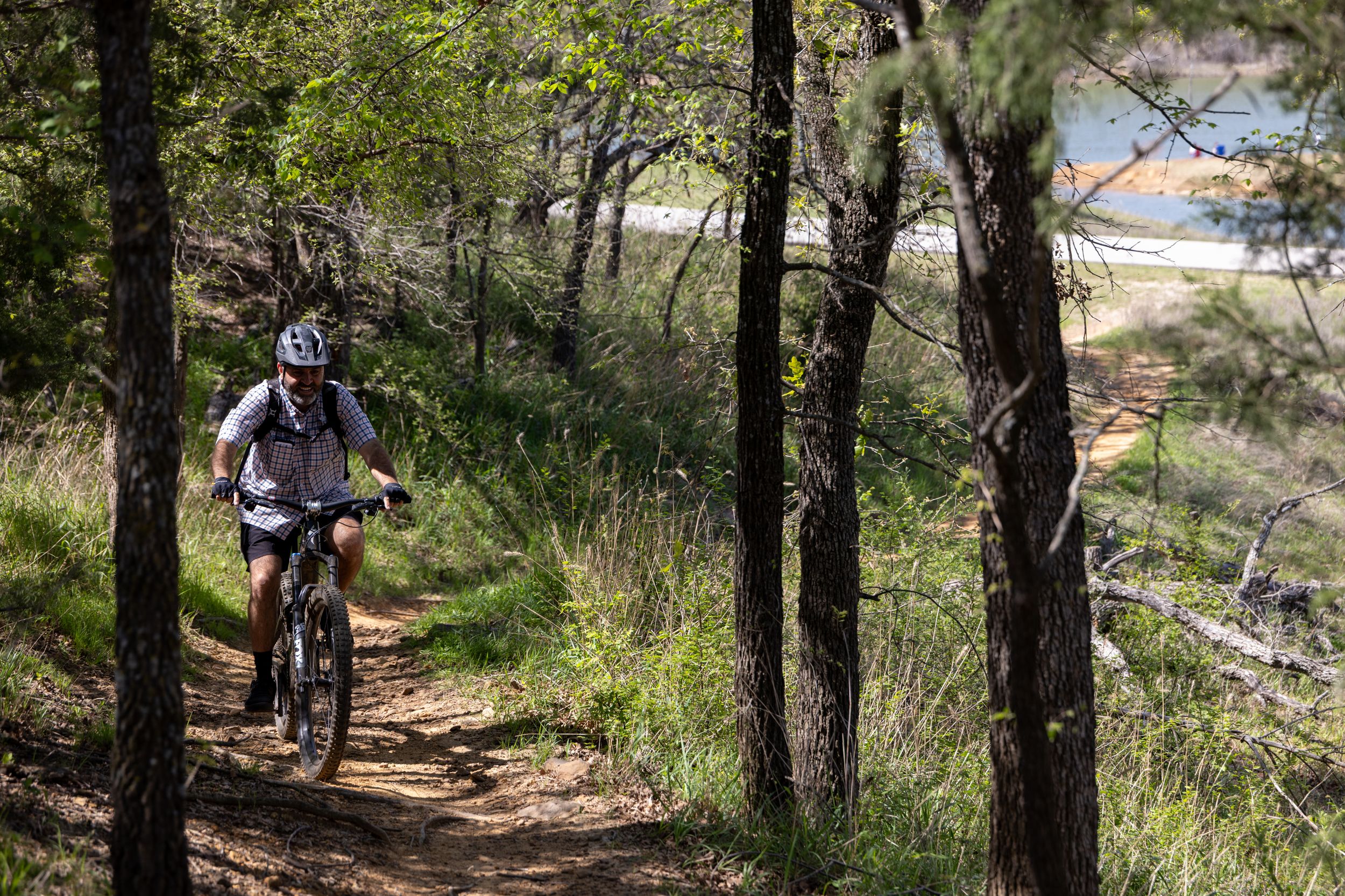 Mountain biker riding a dirt trail through a pine forest near a lake on a sunny day.