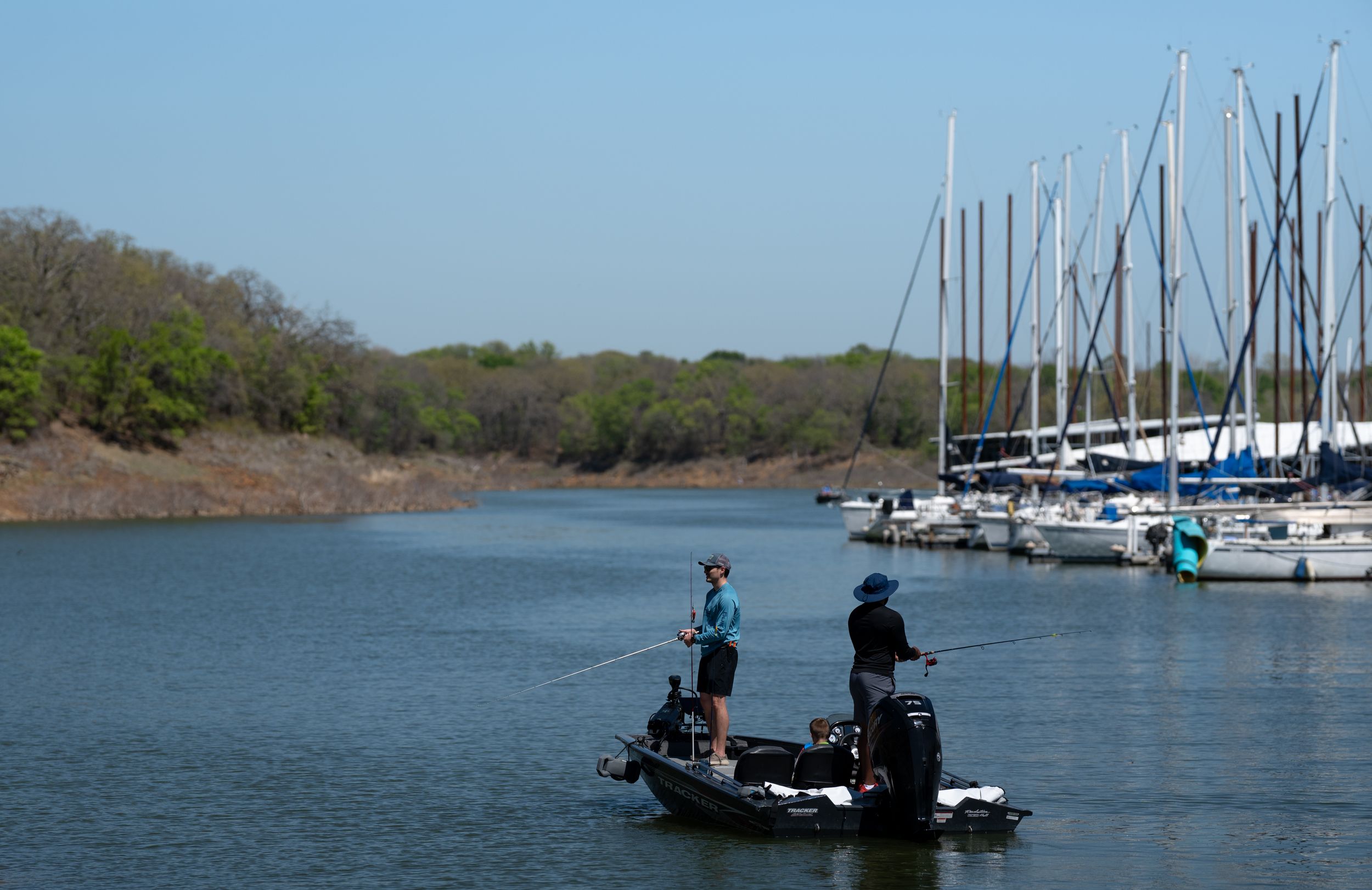 Two anglers fishing from a small boat near a marina with sailboats on a calm lake surrounded by trees.