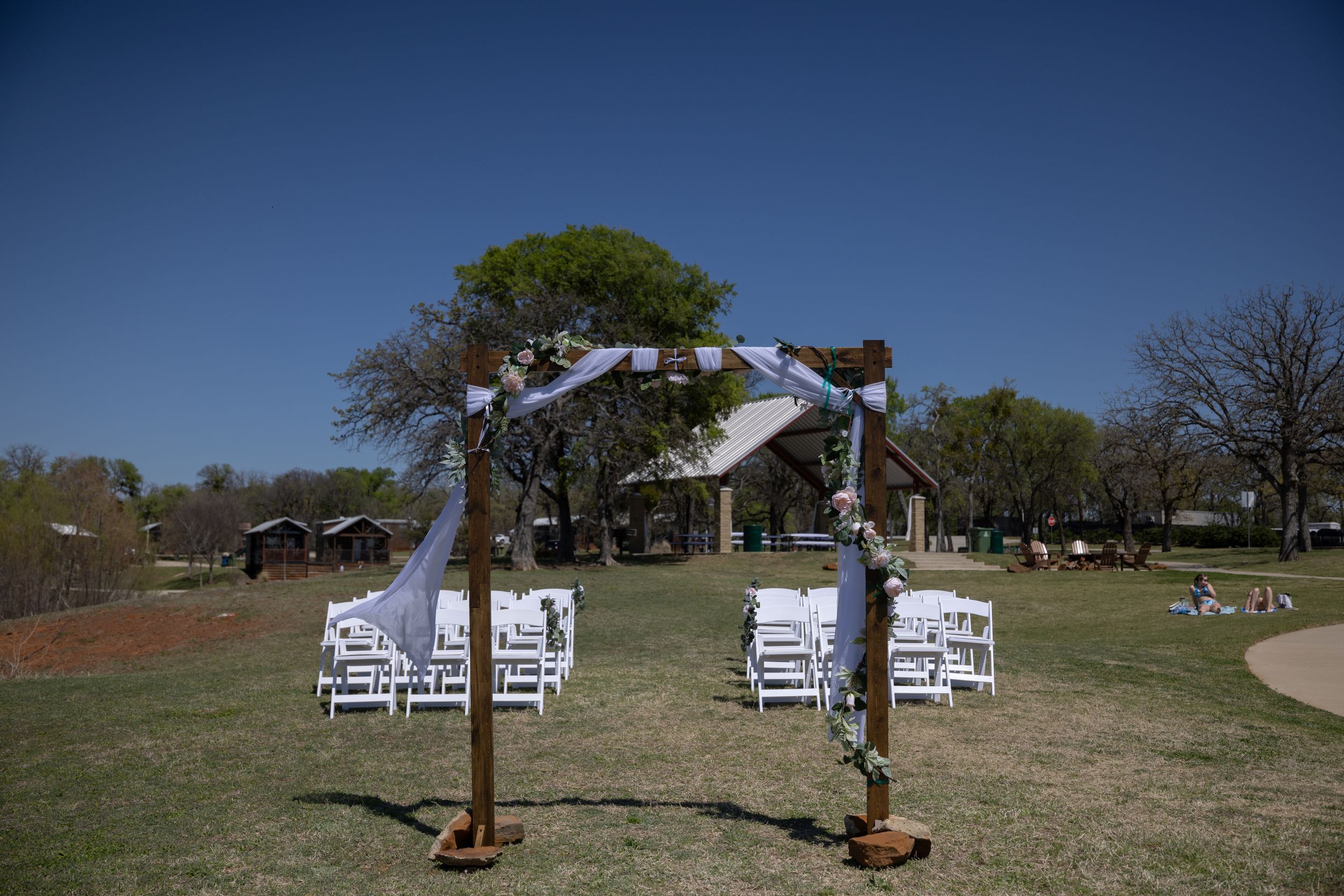 Outdoor wedding ceremony setup with white chairs and wooden arch decorated with fabric on a green lawn.
