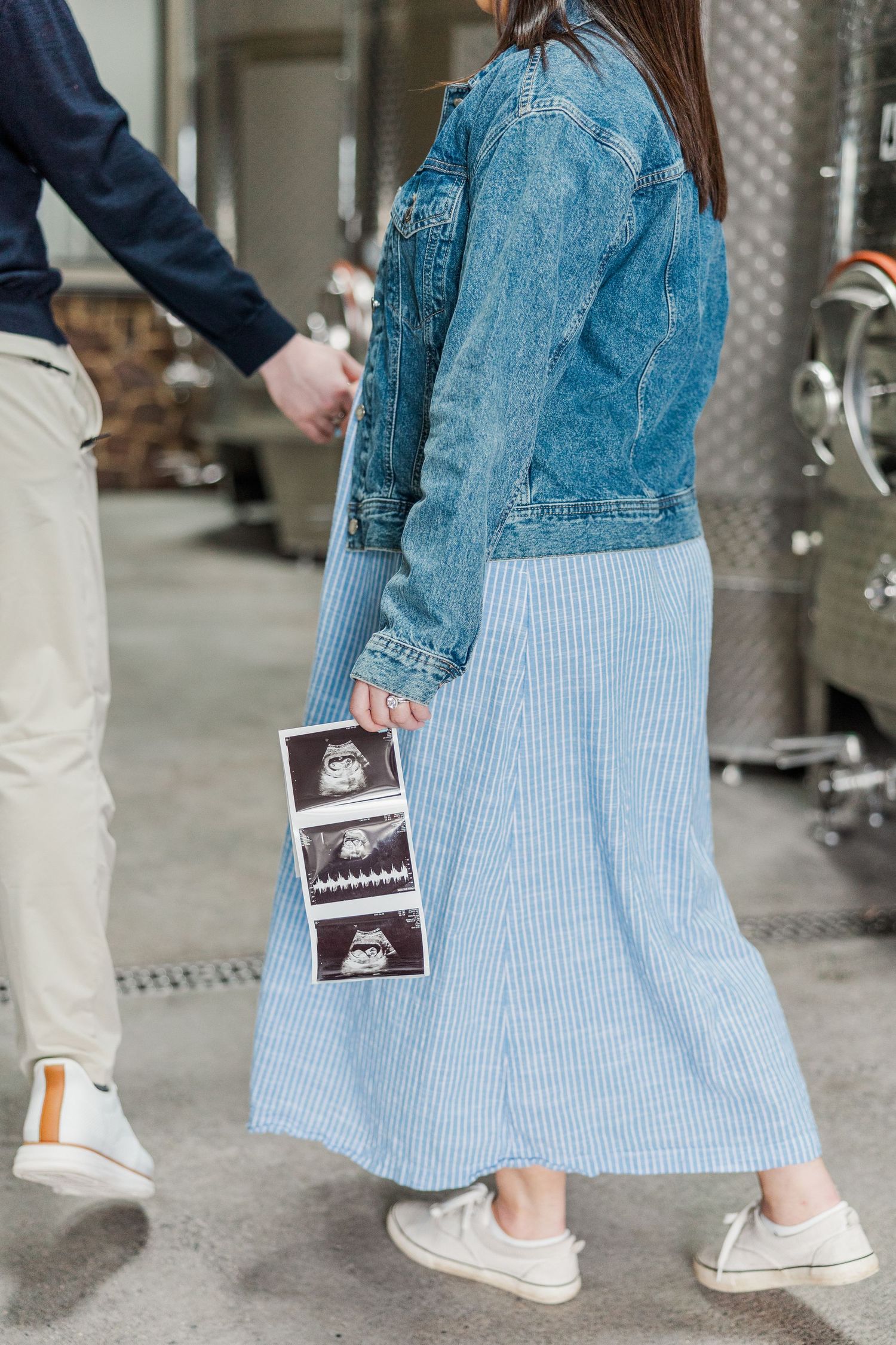 Pregnant woman in denim jacket and striped dress cradling baby bump, smiling couple maternity photo.