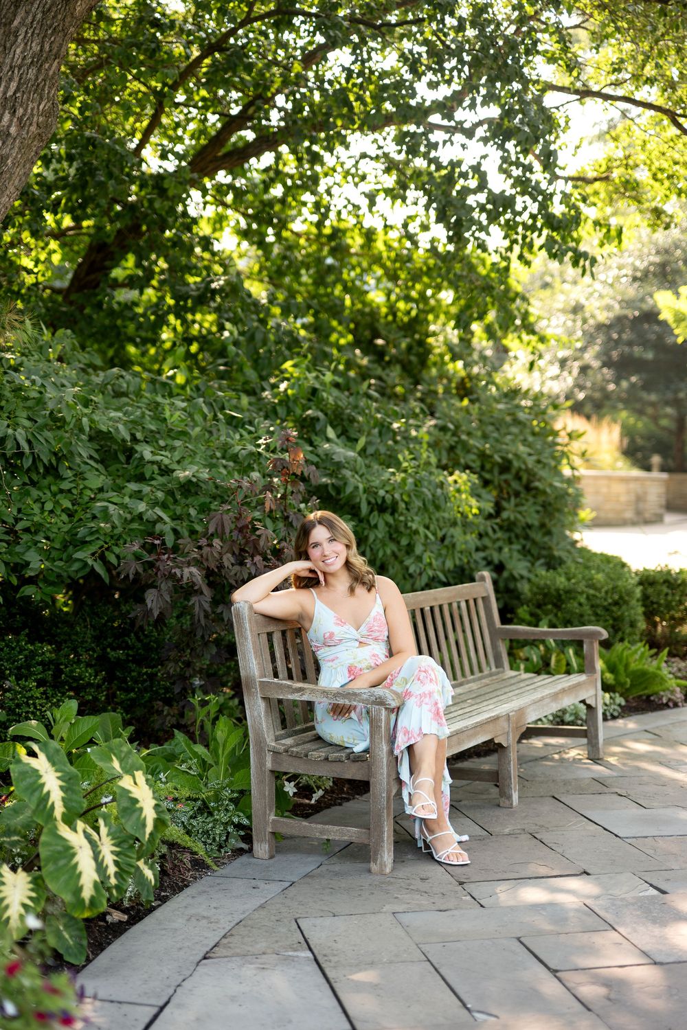 Young woman in floral maxi dress sitting relaxed on wooden garden bench surrounded by lush greenery.
