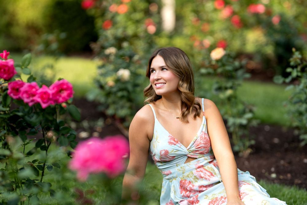 Woman in floral dress smiling among vibrant pink roses in a lush garden setting.