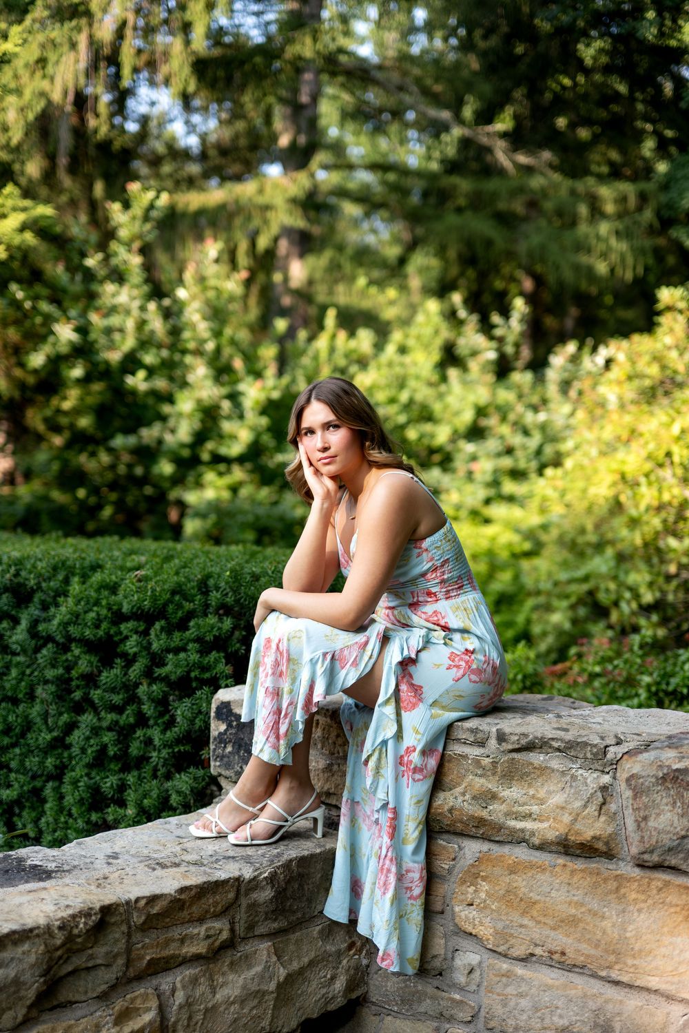 Young woman in floral jumpsuit sitting thoughtfully on stone wall surrounded by green garden foliage.