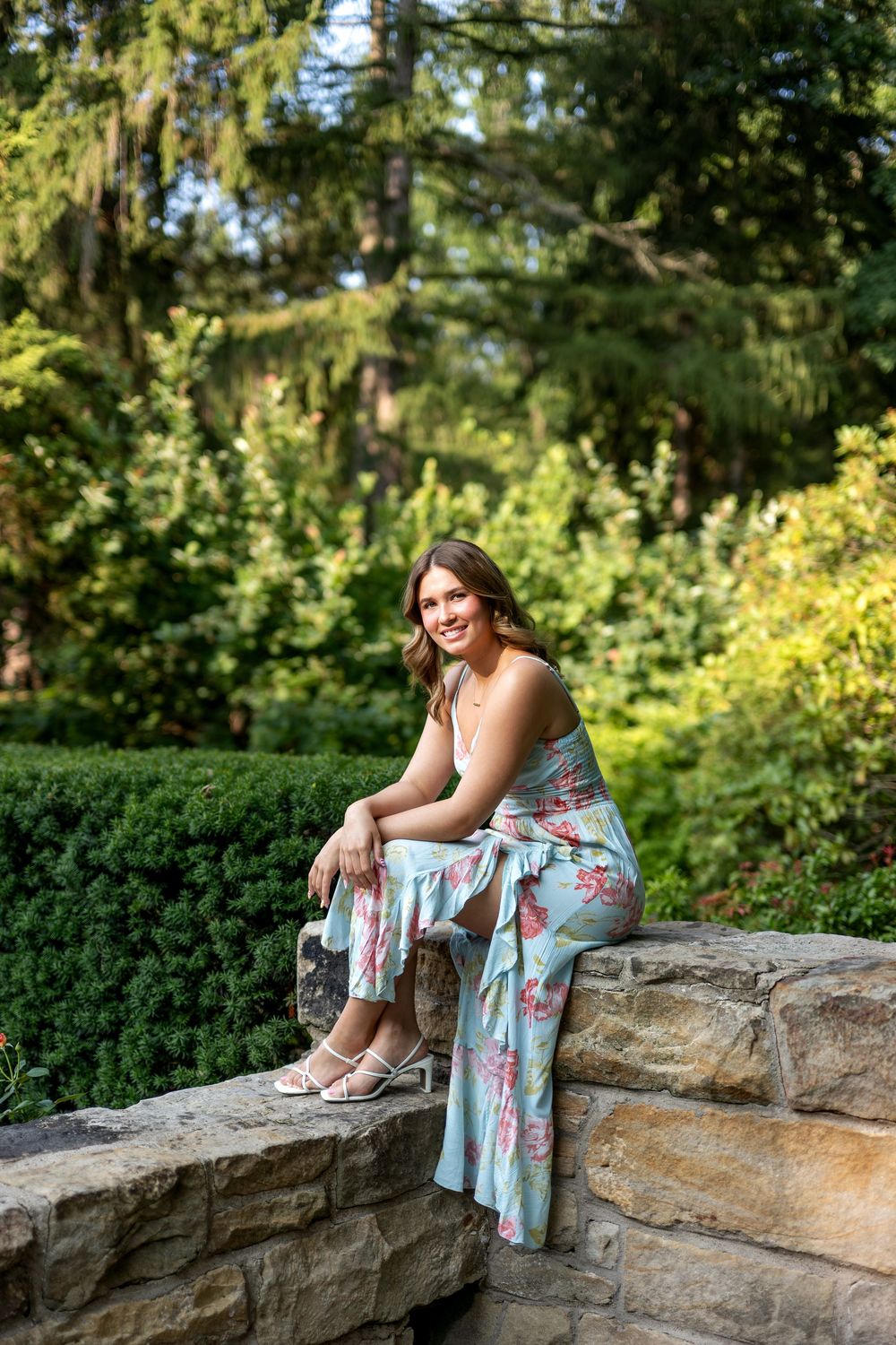 Young woman in floral mint jumpsuit sitting on stone wall surrounded by lush green garden trees.