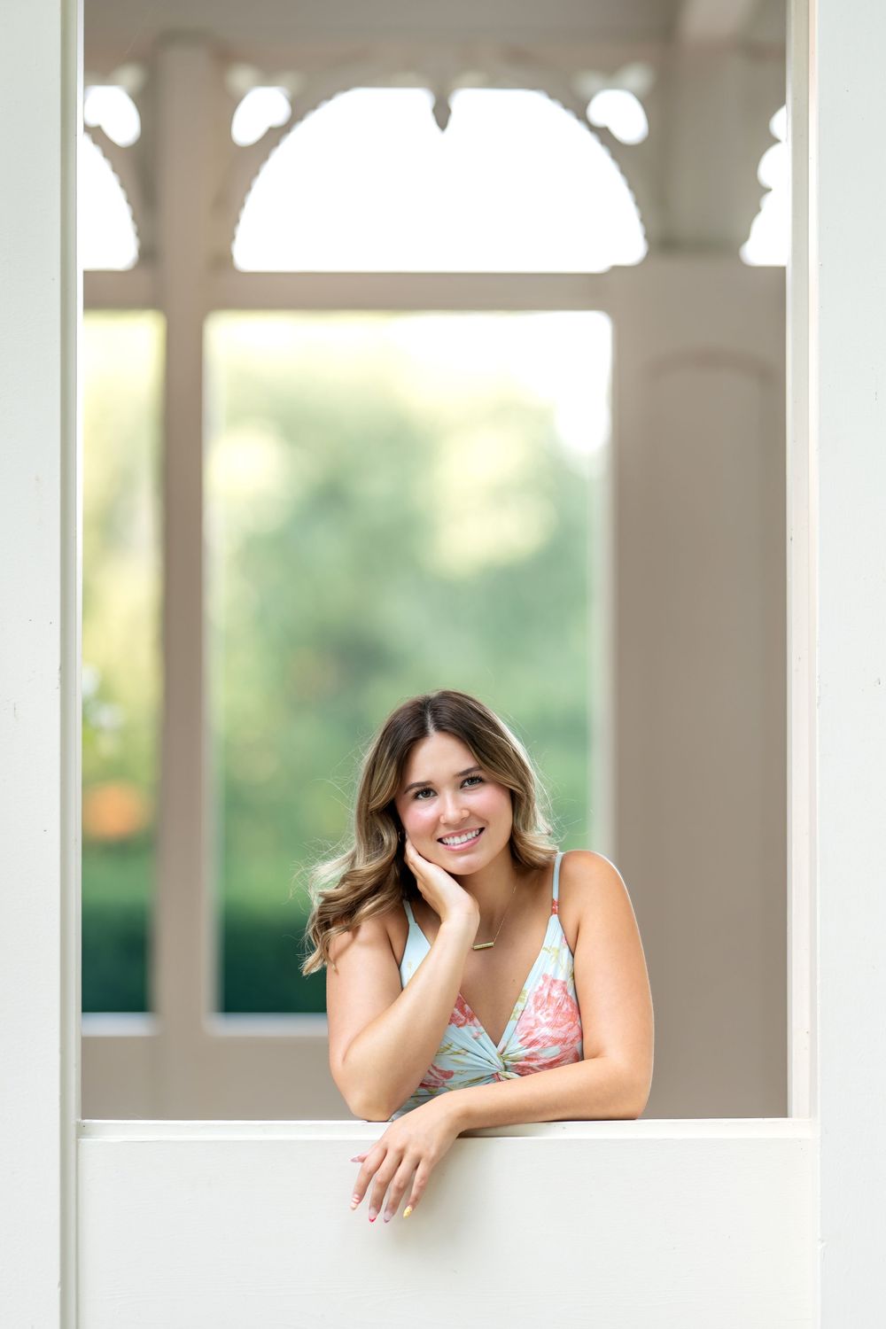 Smiling young woman in floral dress leaning on white gazebo railing with garden backdrop.