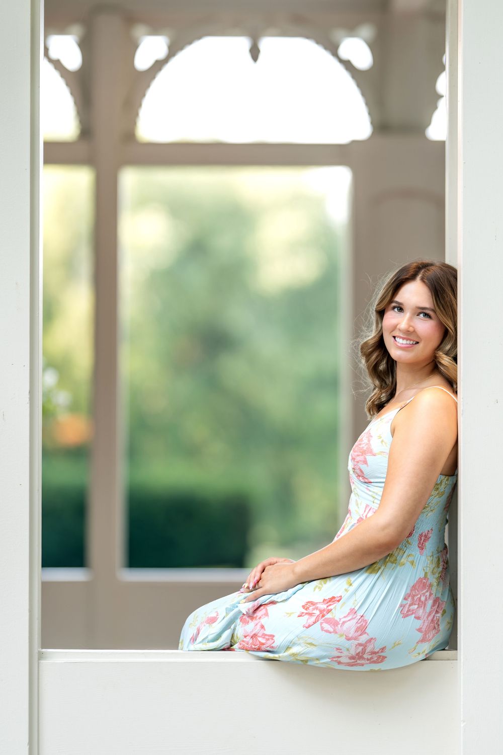 Cheerful young woman in blue floral dress seated inside elegant white garden gazebo structure.