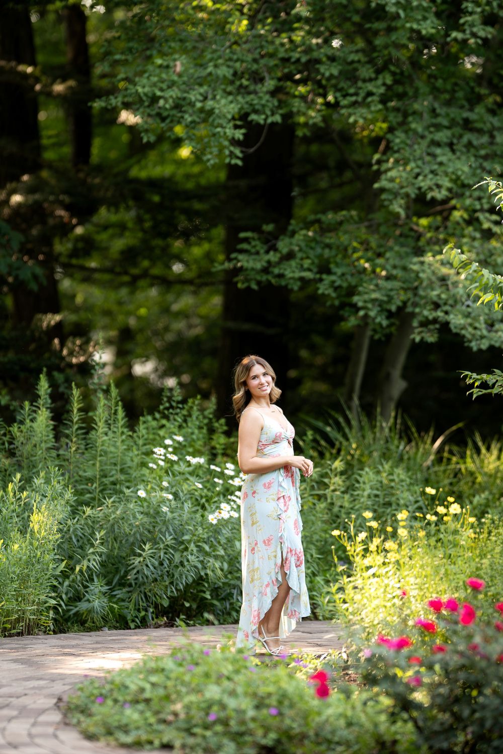 Woman in floral maxi dress standing on garden path surrounded by lush greenery and colorful flowers.