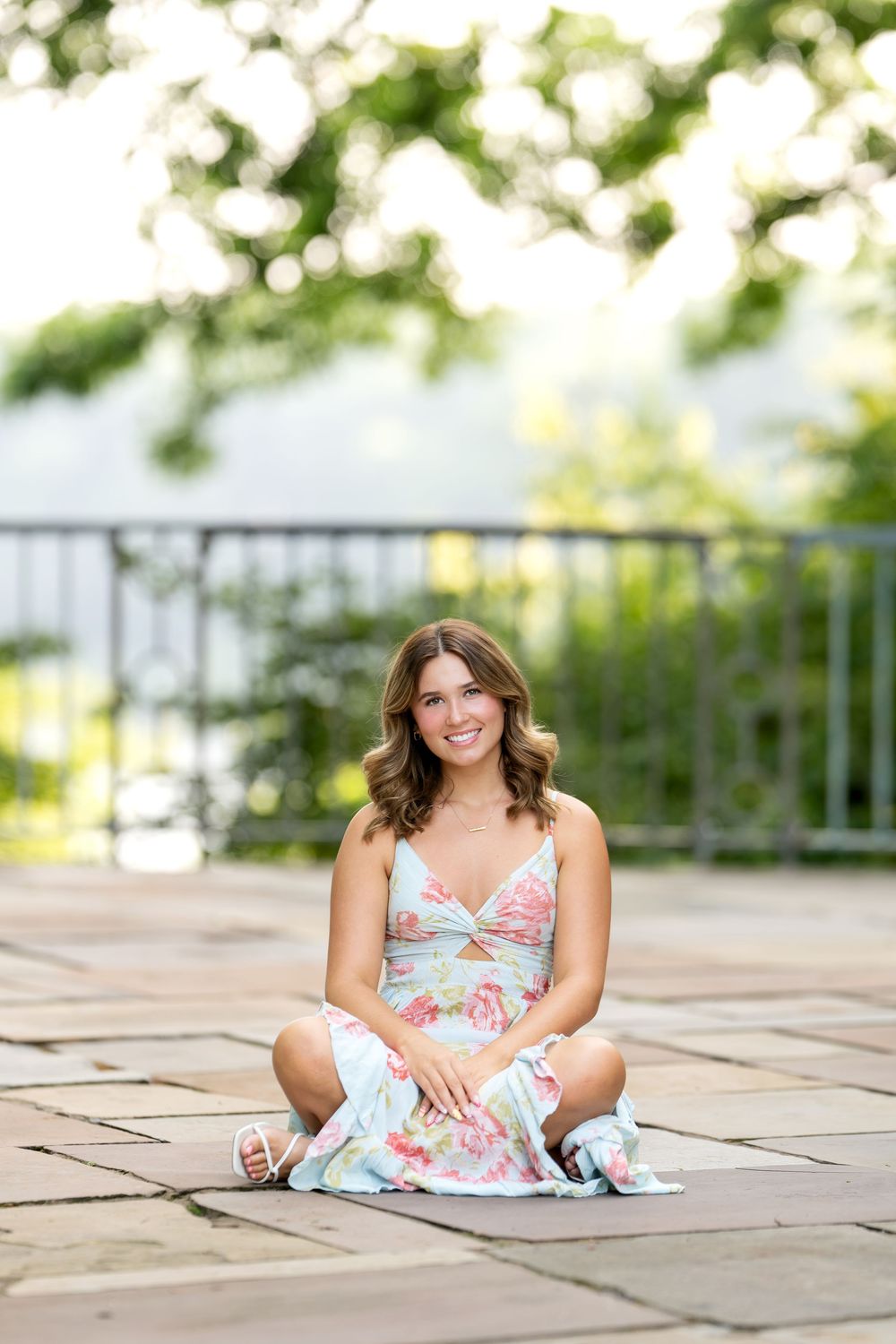 Young woman in floral sundress sitting cross-legged on stone patio surrounded by lush greenery.