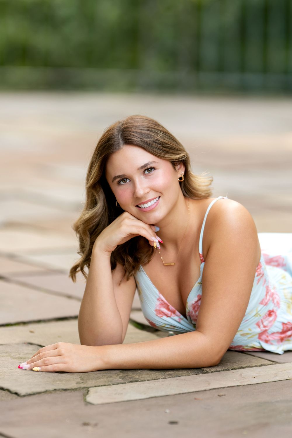 Young woman in floral sundress lying on brick patio, smiling warmly with hand on chin, outdoors.