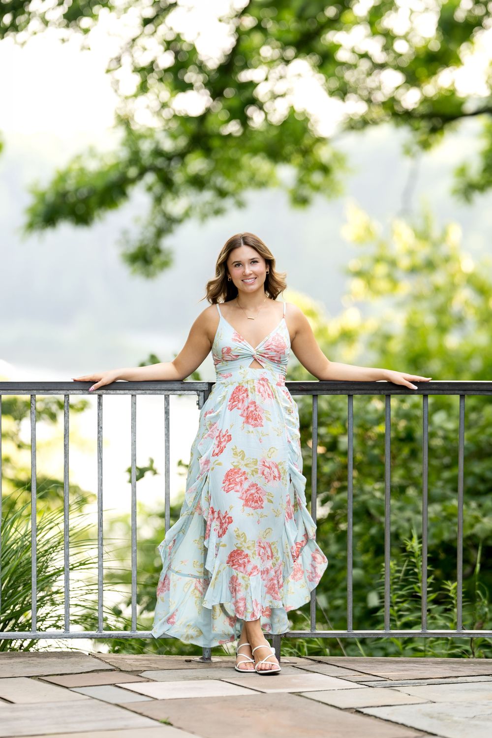 Smiling young woman in blue floral maxi dress leaning on metal railing with lush green backdrop.