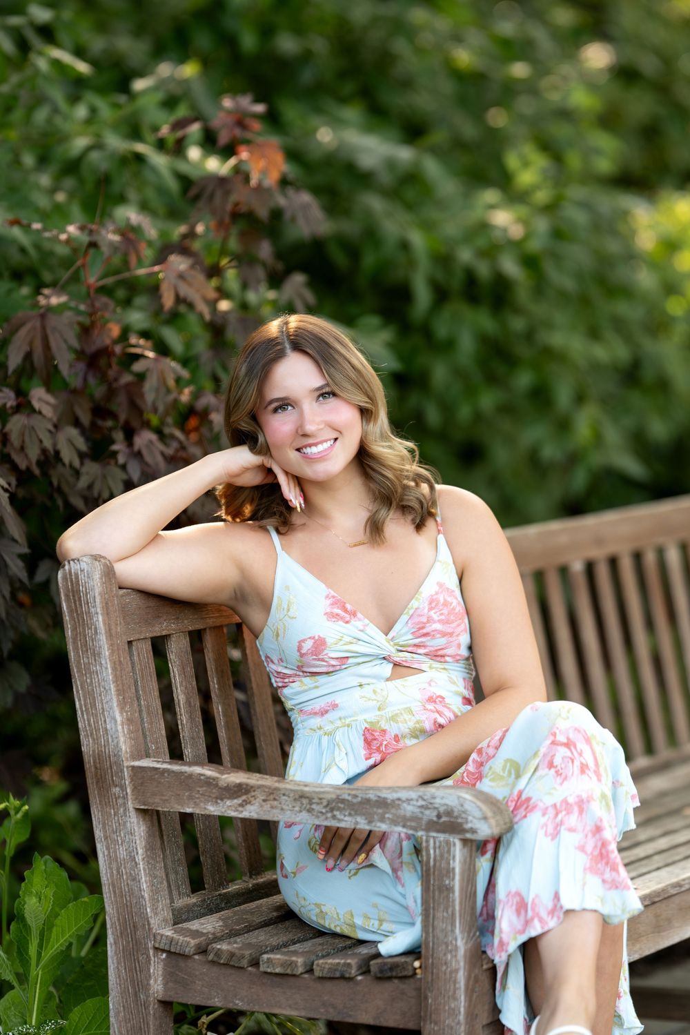 Young woman in floral sundress sitting on wooden bench in lush garden, smiling cheerfully.