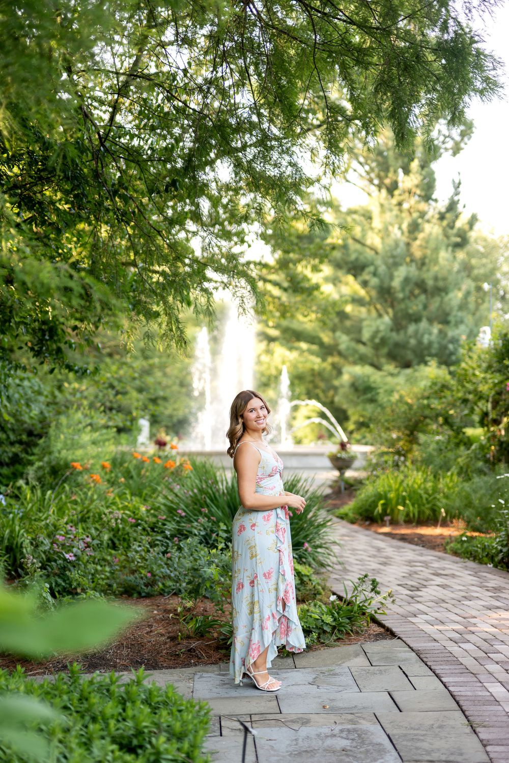 A woman in a floral maxi dress poses on a garden path surrounded by lush greenery and a fountain.
