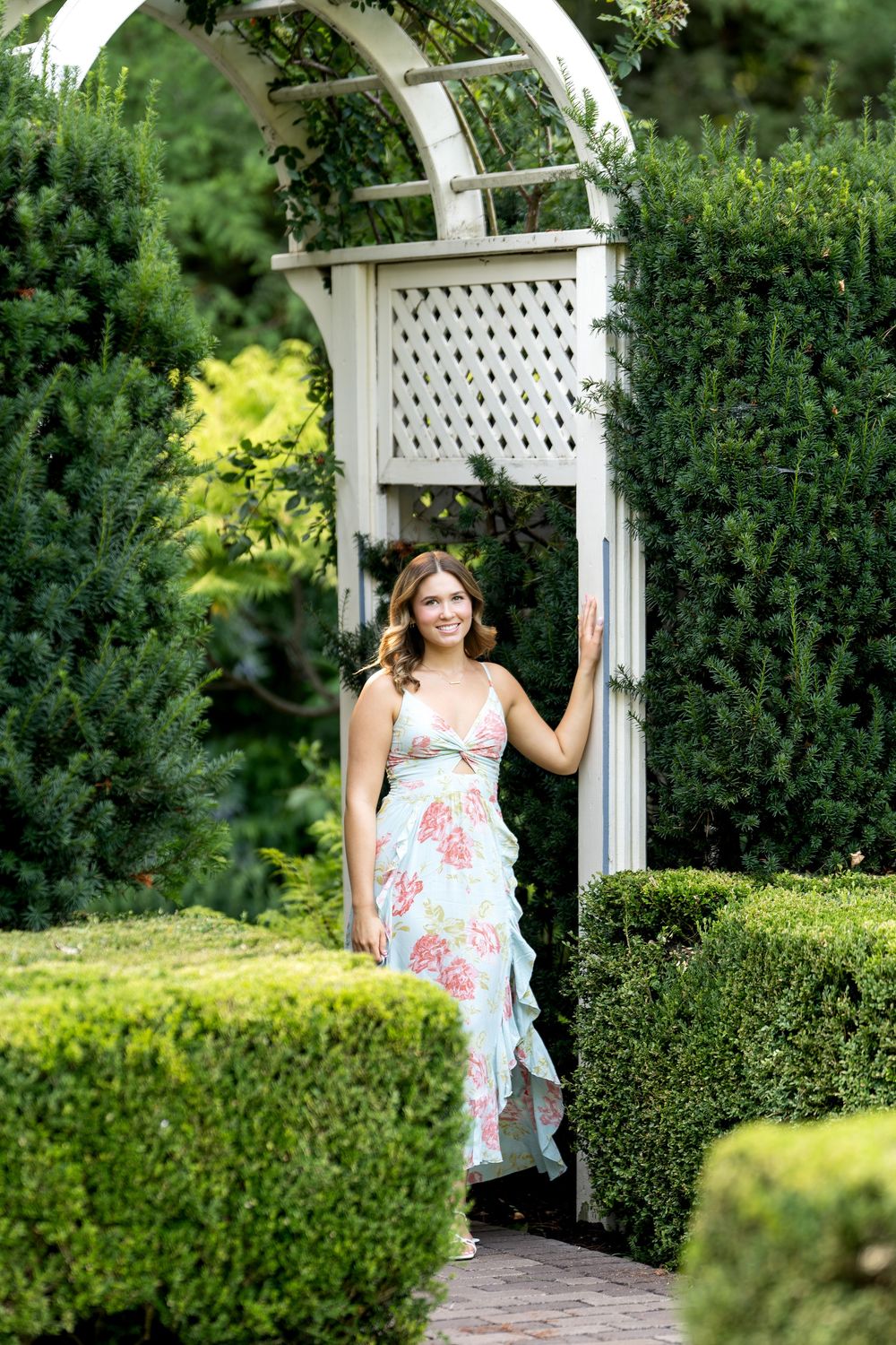 Woman in floral midi dress standing under white garden arbor surrounded by lush green hedges.