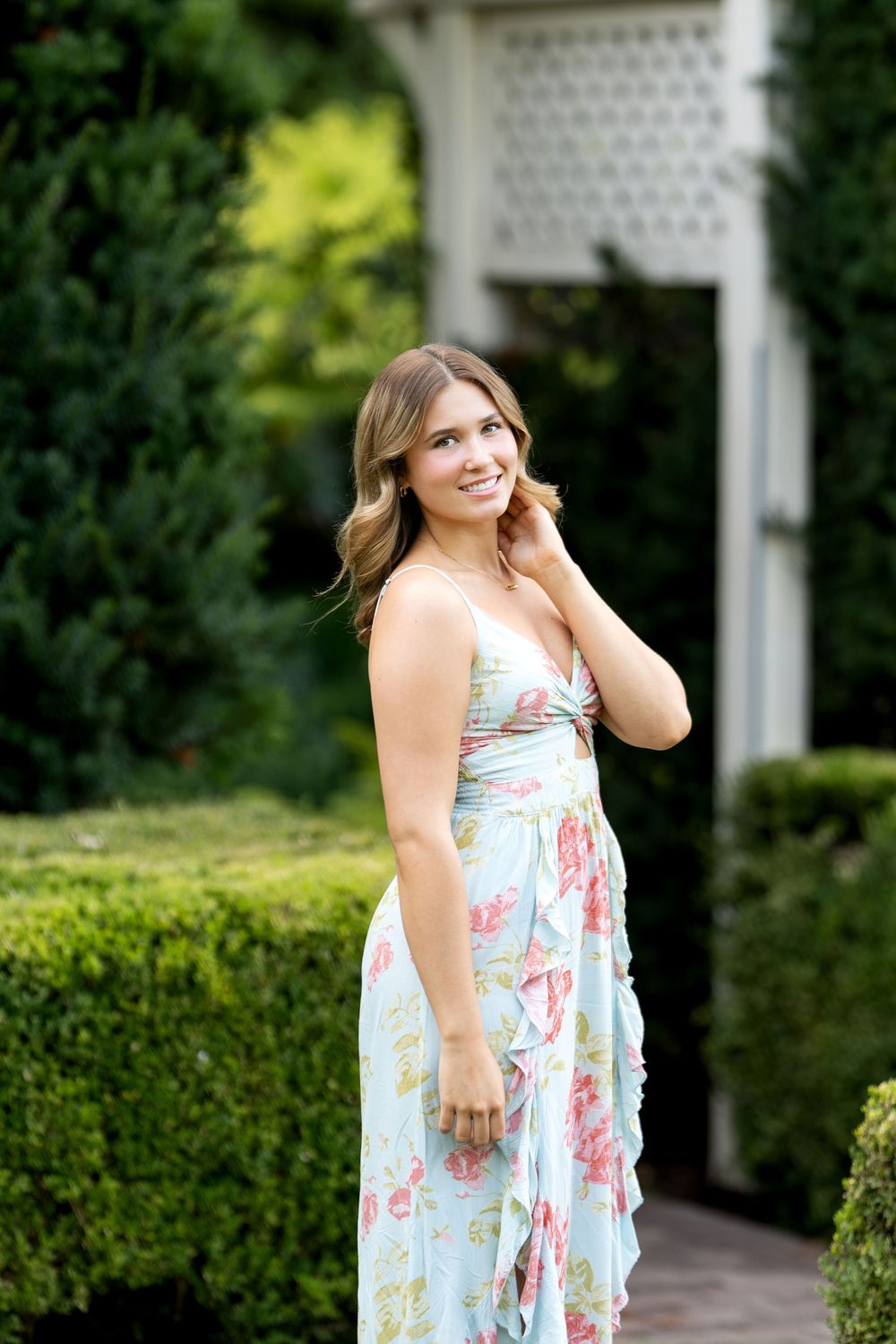 Young woman in a light blue floral maxi dress smiling in a lush green garden with hedges and white arbor.