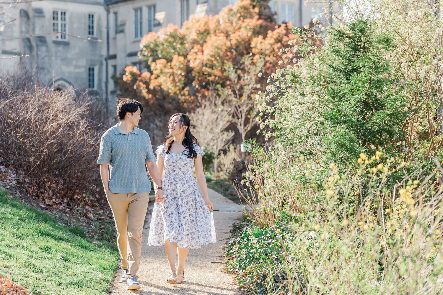 Couple walking hand in hand along a garden path beside a historic building with colorful autumn foliage.