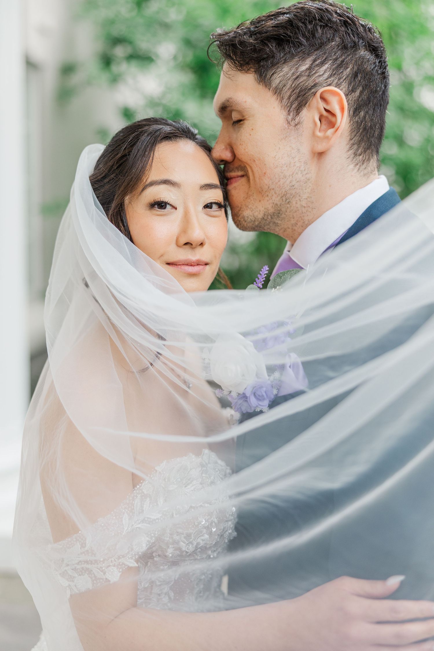 A bride and groom share an intimate moment wrapped in a flowing white veil during their wedding day portrait.