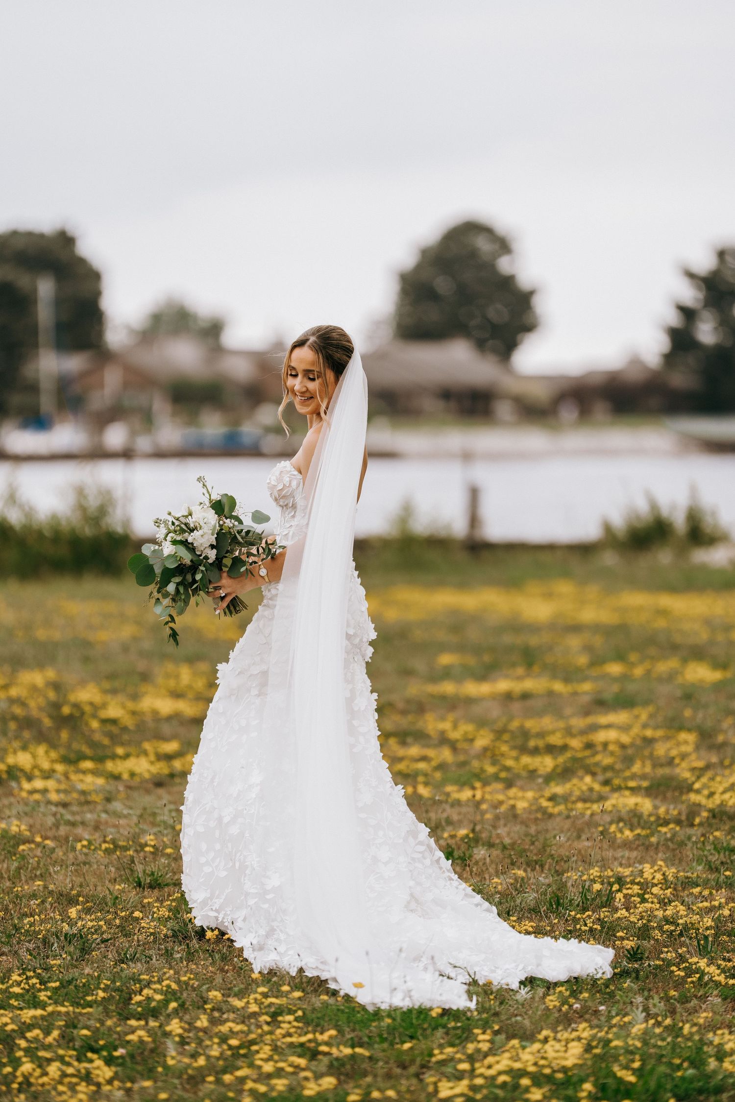Bride in white floral gown with veil holds bouquet in yellow wildflower field near waterfront.