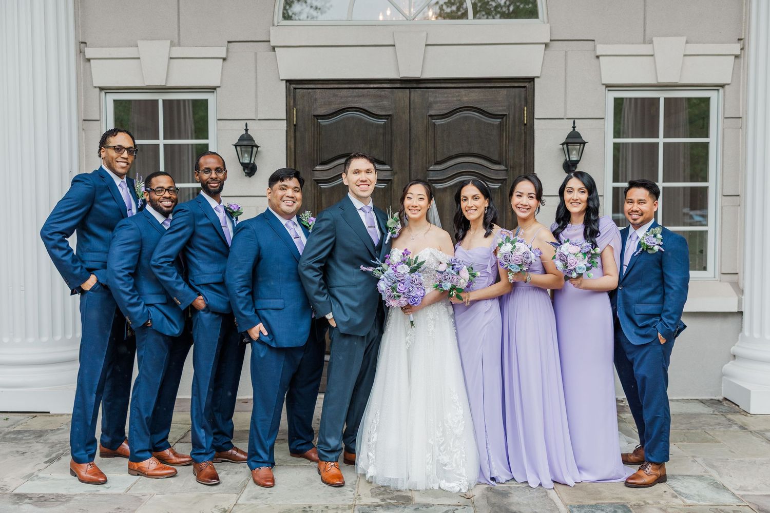 Wedding party poses outdoors, groomsmen in navy suits and bridesmaids in lavender dresses holding floral bouquets.