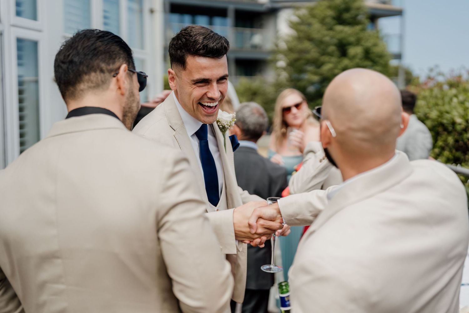 Groom in beige suit shaking hands with guests at outdoor wedding reception on sunny day.