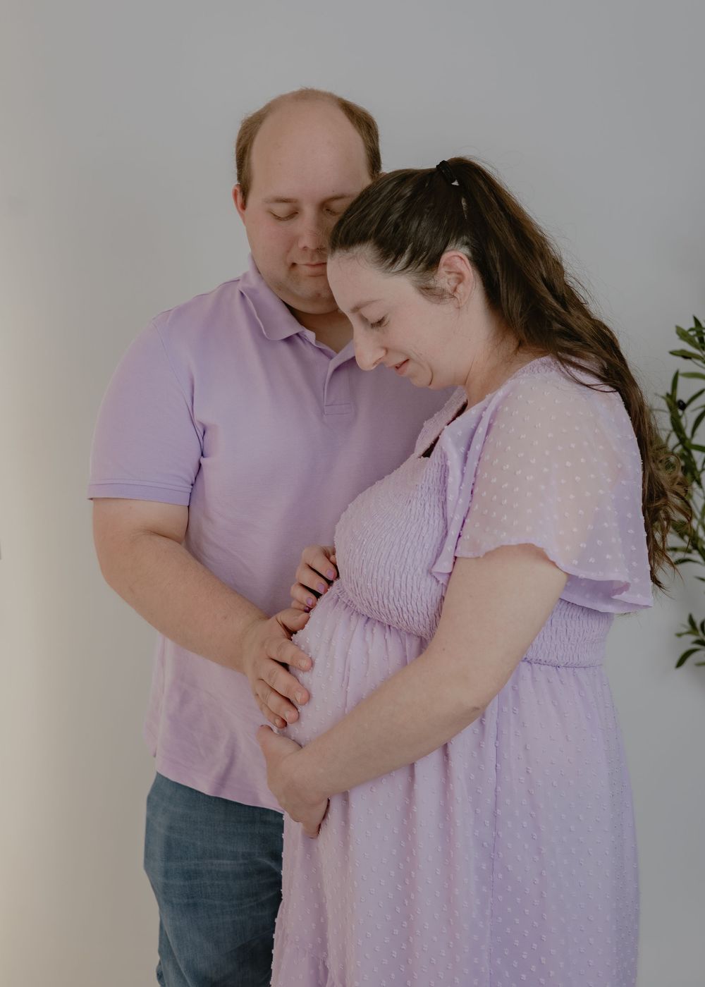Couple tenderly holds pregnant belly together in a soft lavender and white studio maternity portrait.