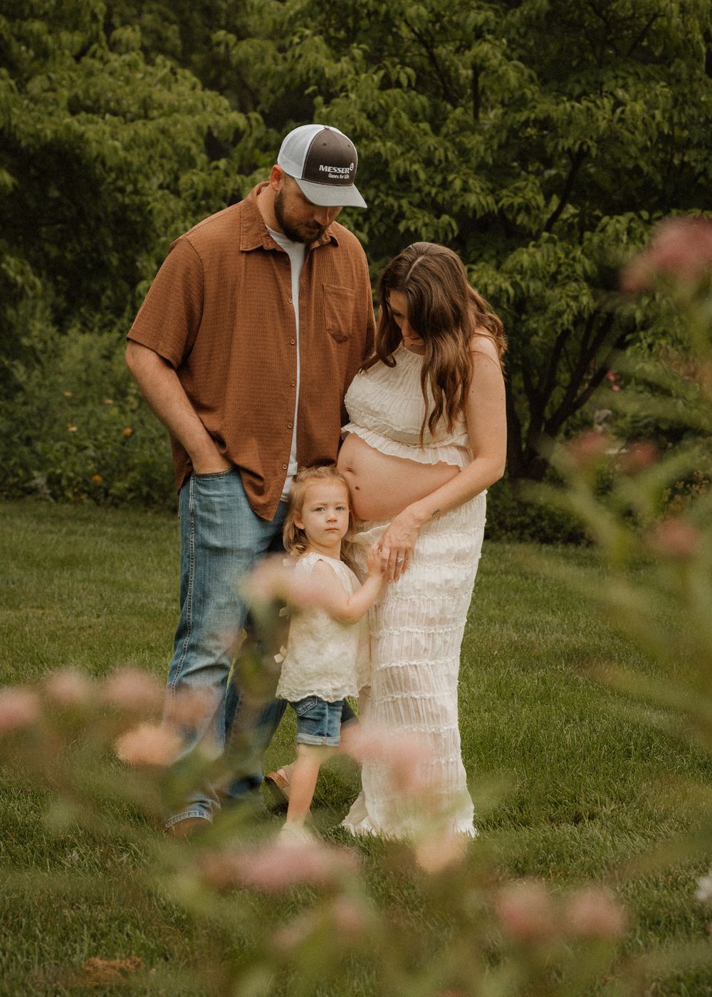 Pregnant woman in lace dress with husband and toddler standing in lush green garden setting.
