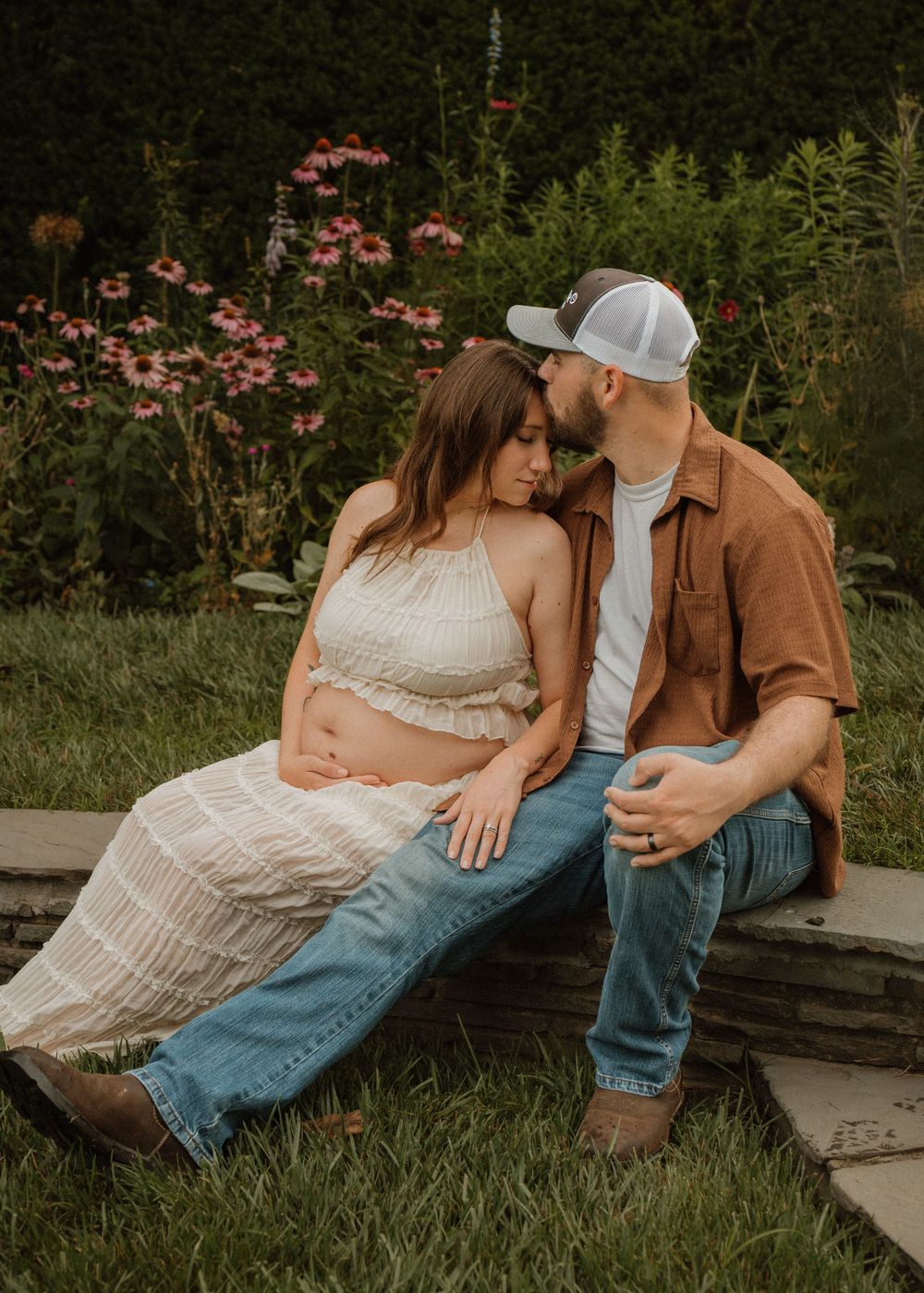 Couple sharing tender moment outdoors, man kissing pregnant womans head while seated in garden.