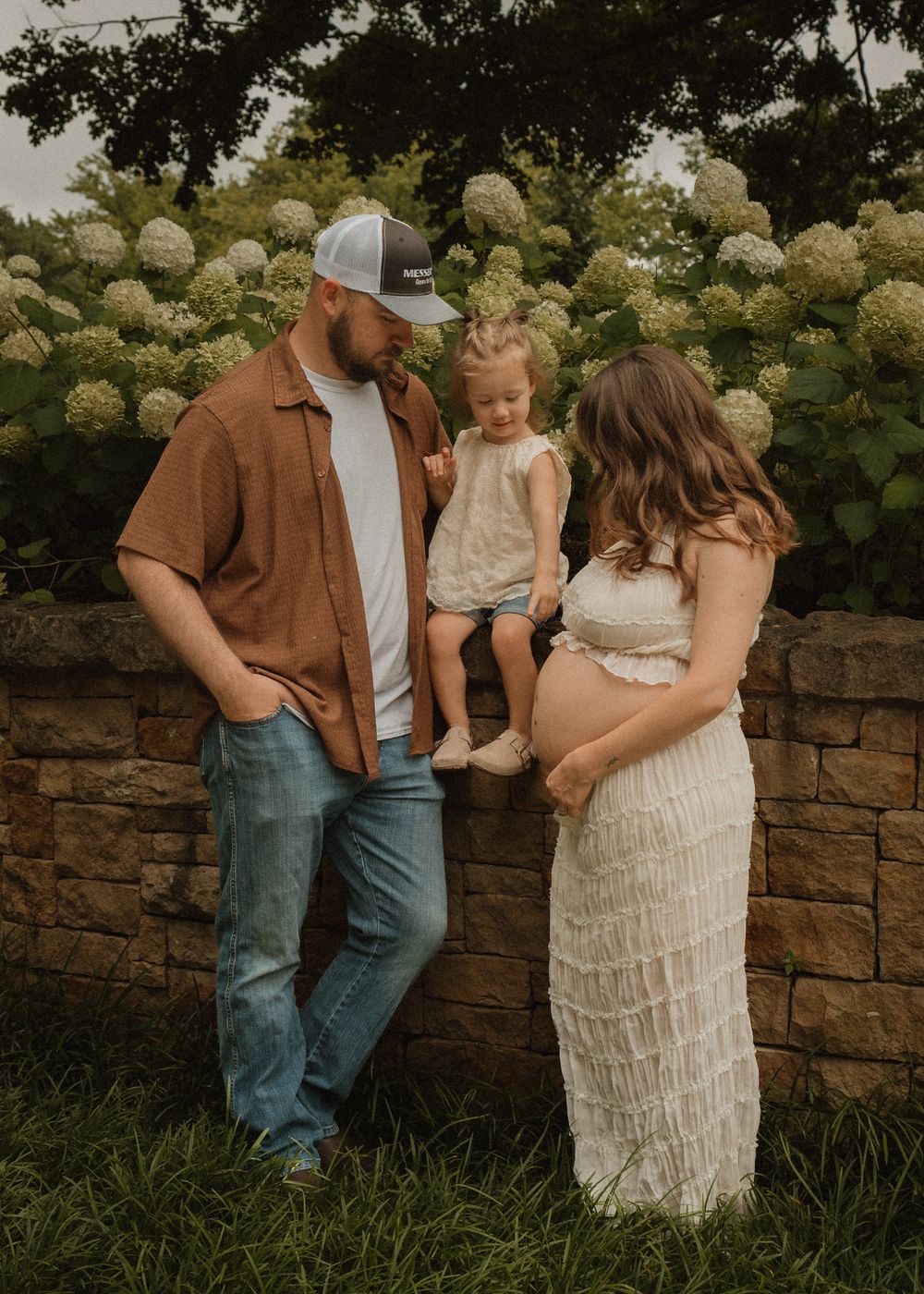 Expecting couple with toddler posing by stone wall with white hydrangeas in outdoor garden.