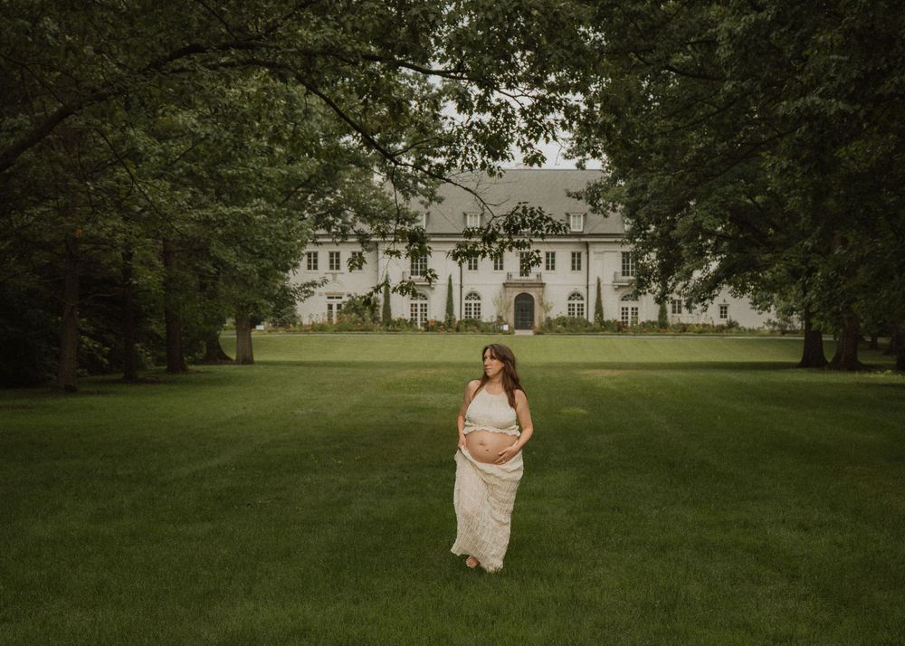 Pregnant woman in flowing white dress standing on vast green lawn before elegant white estate.