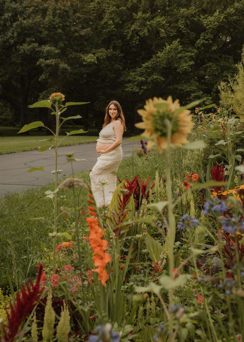Pregnant woman in cream outfit standing among vibrant colorful wildflowers in a lush garden.