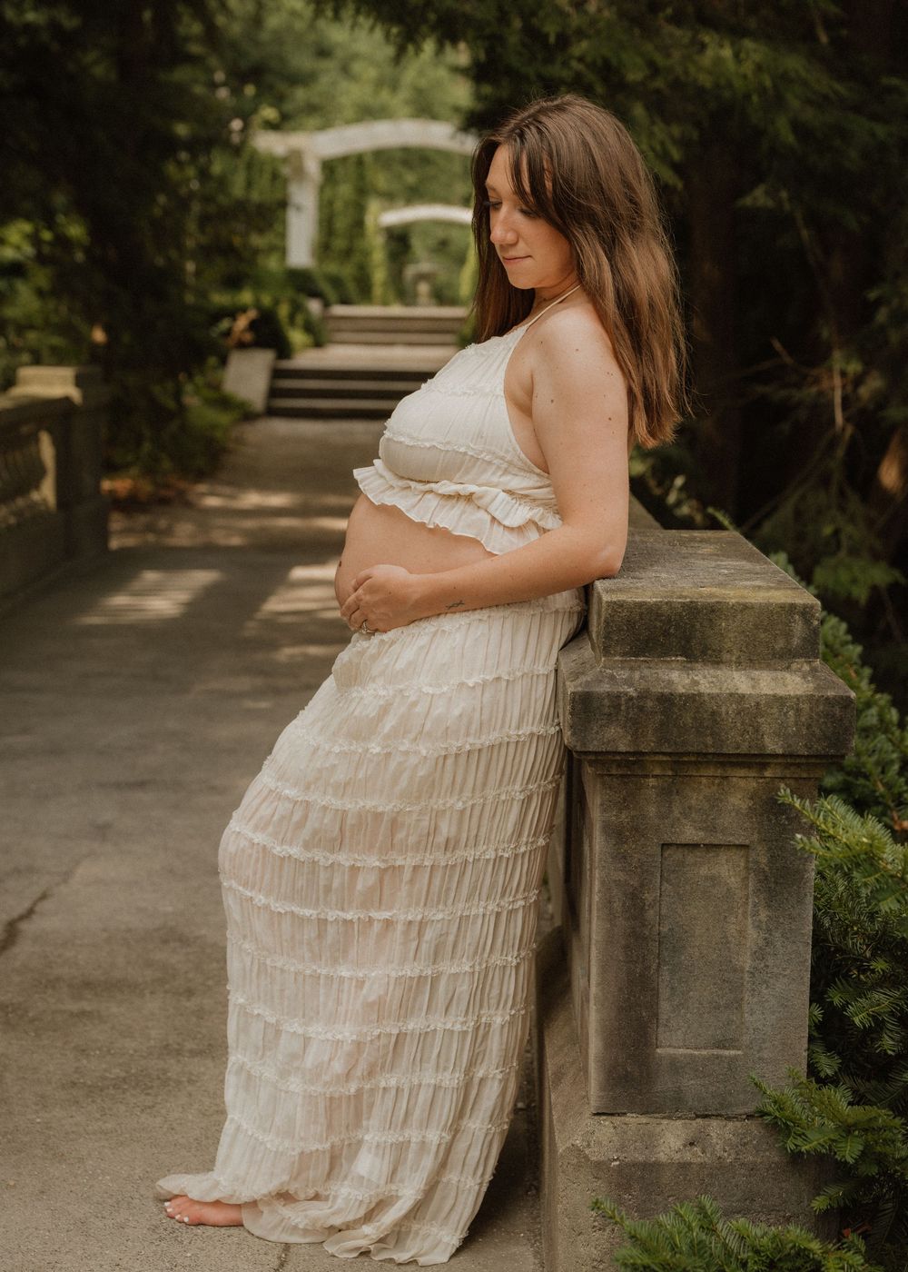 Pregnant woman in white ruffled crop top and maxi skirt poses gracefully in a lush garden setting.