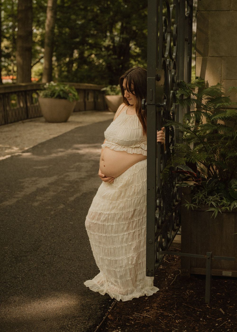 Expectant mother in elegant white tiered dress stands by a garden gate, cradling her baby bump.