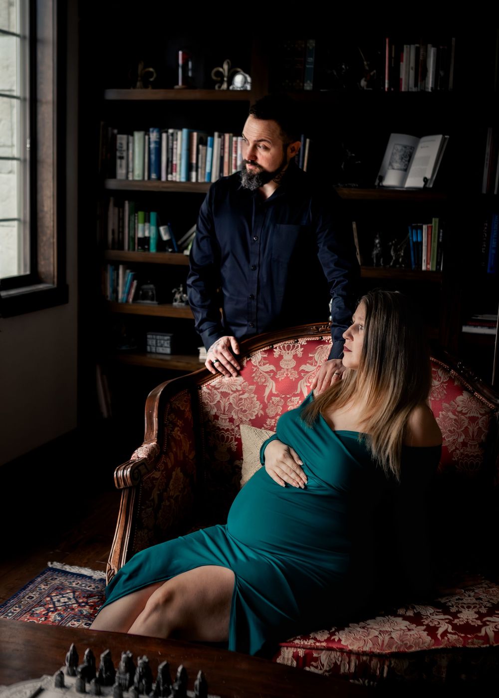 Pregnant woman in teal off-shoulder gown reclines in ornate chair beside partner in a moody library.