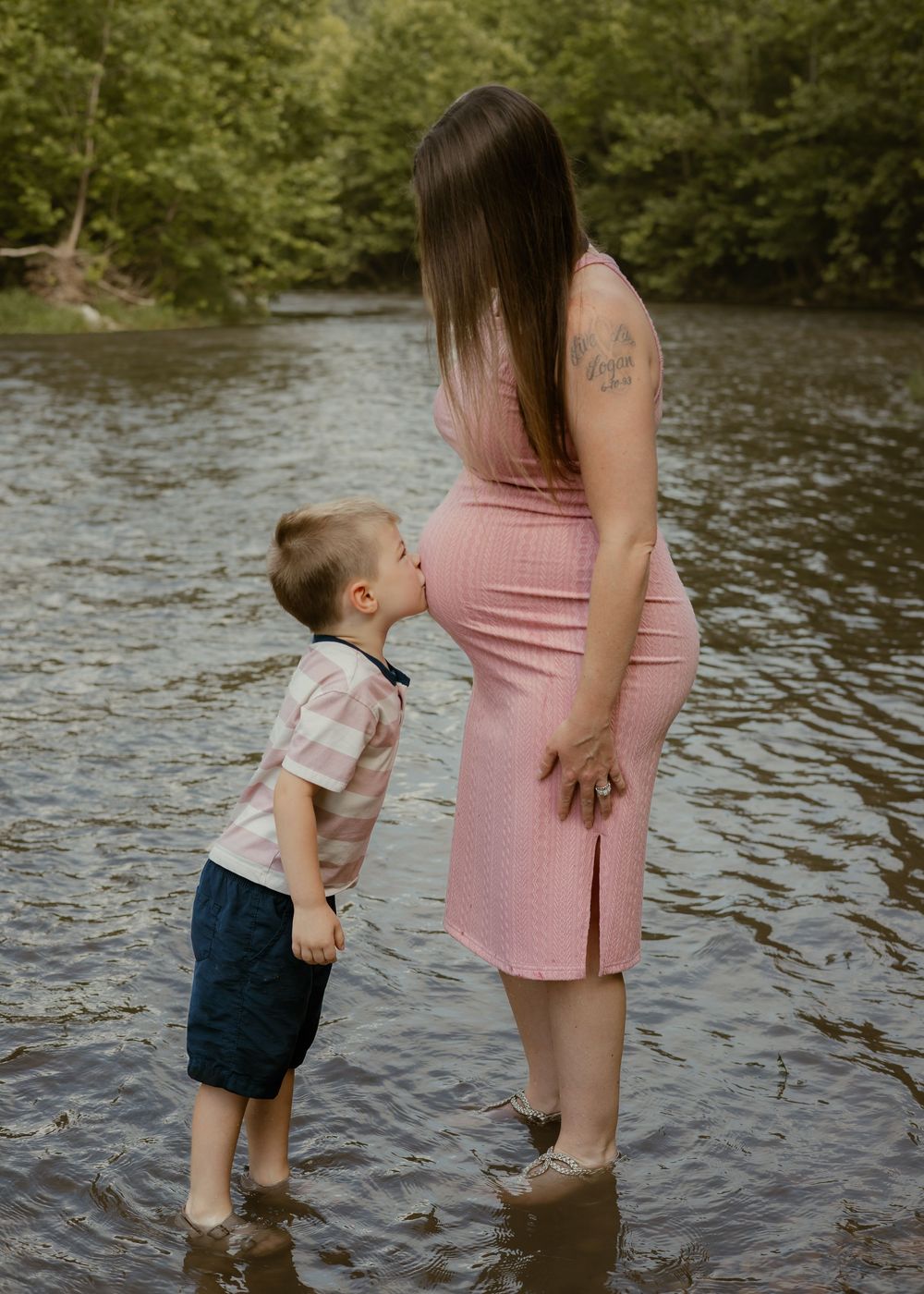 Young boy sweetly kisses his pregnant mother's baby bump while standing in a shallow river.