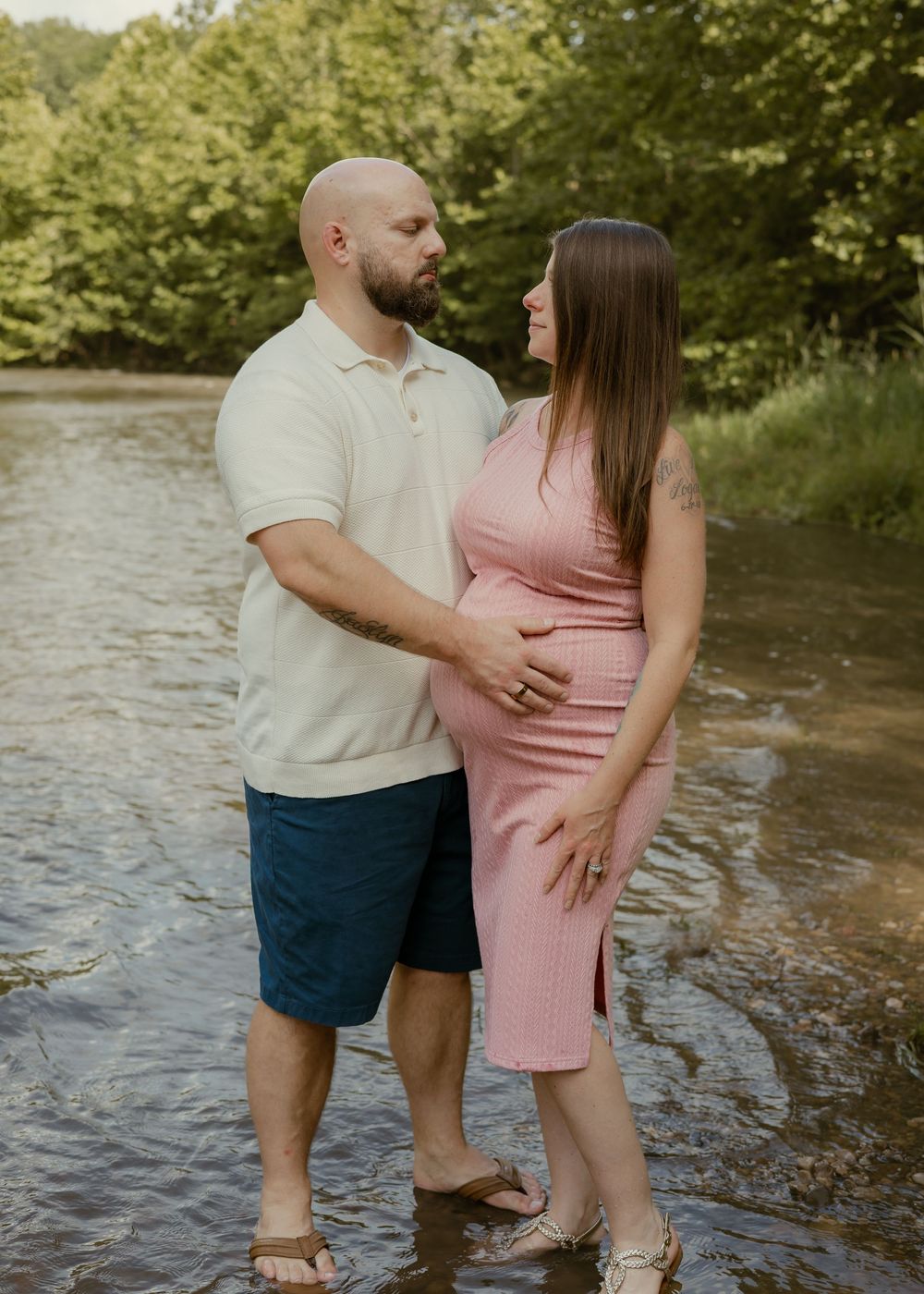 Expecting couple shares a tender moment together standing in a peaceful river surrounded by trees.