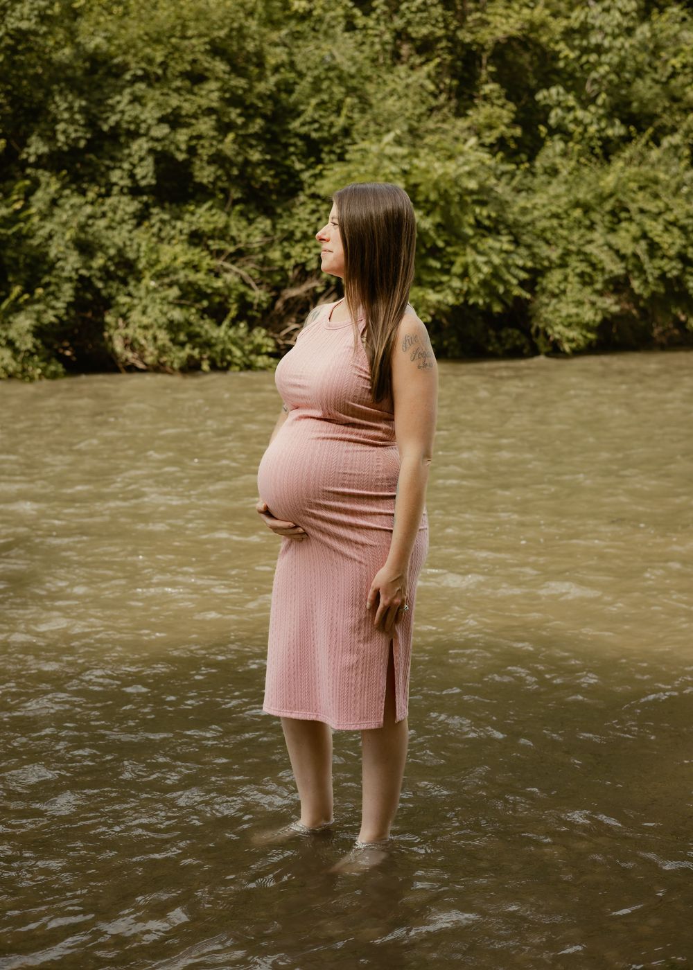 Pregnant woman in pink ribbed dress stands serenely in a river, gently holding her baby bump.