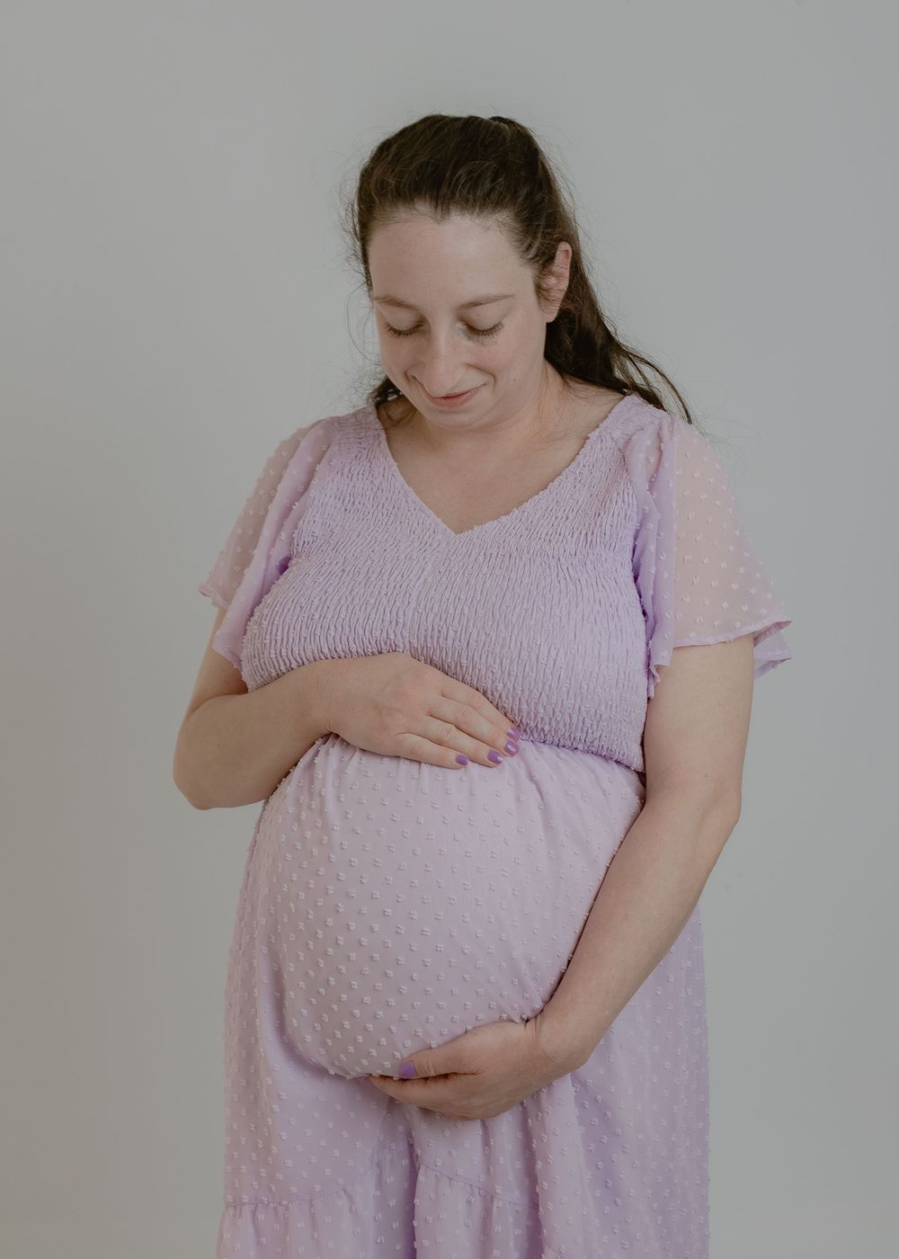 Pregnant woman in lavender flutter-sleeve dress lovingly cradles her baby bump against a neutral background.