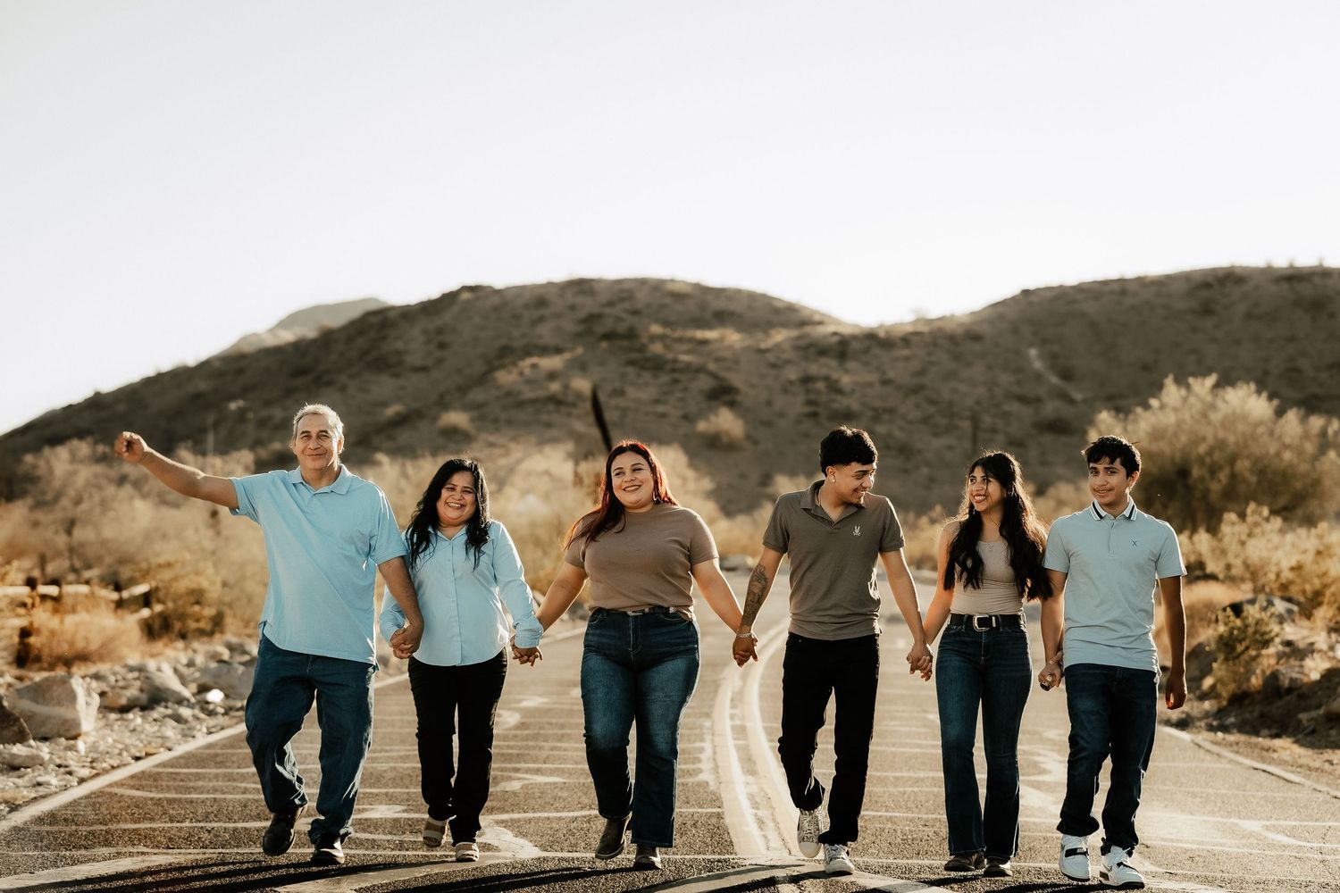 Six family members walk hand-in-hand on a desert road with mountains in the background on a sunny day.
