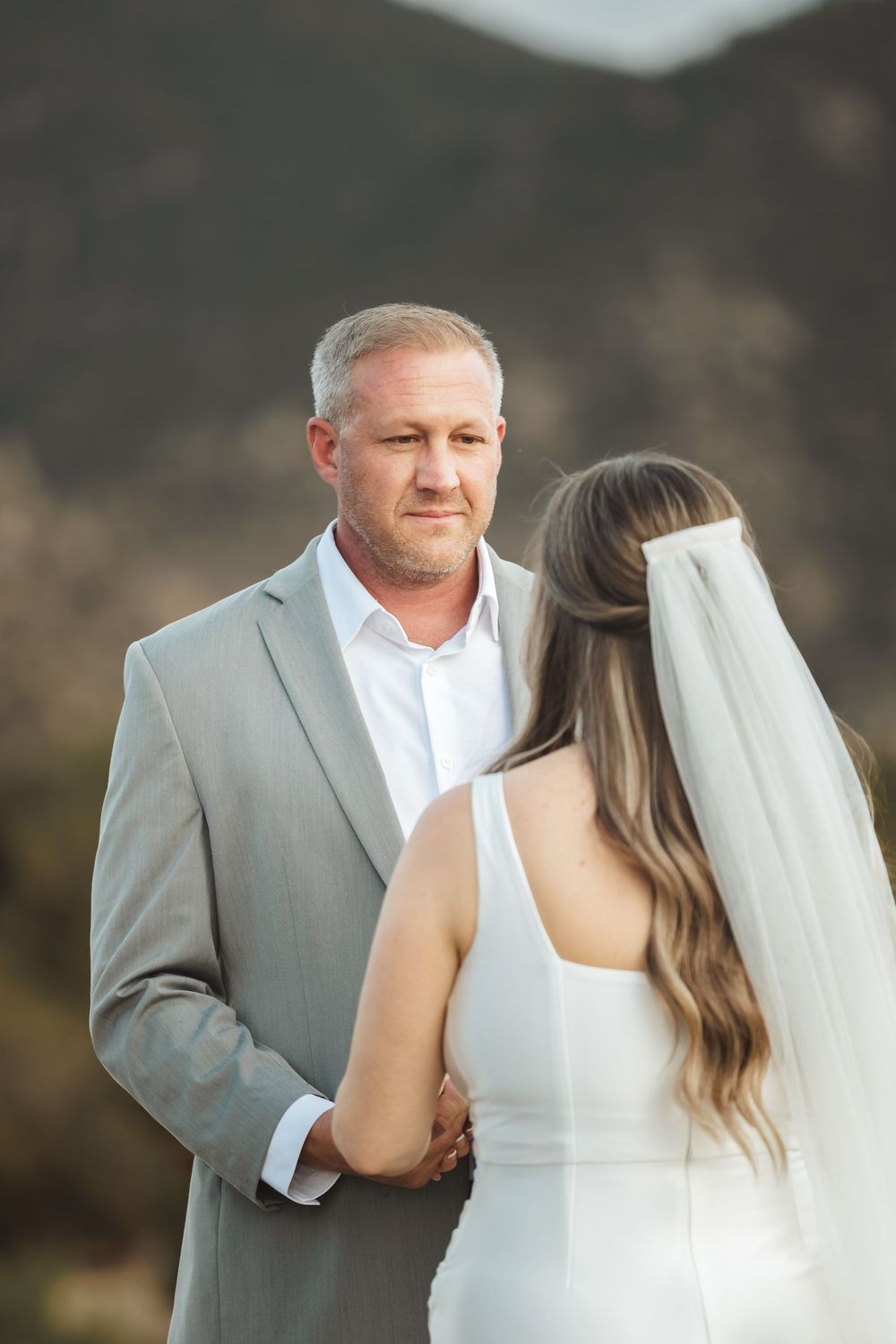 A groom in a gray suit holds hands with a bride in a white dress and veil during an outdoor wedding ceremony.