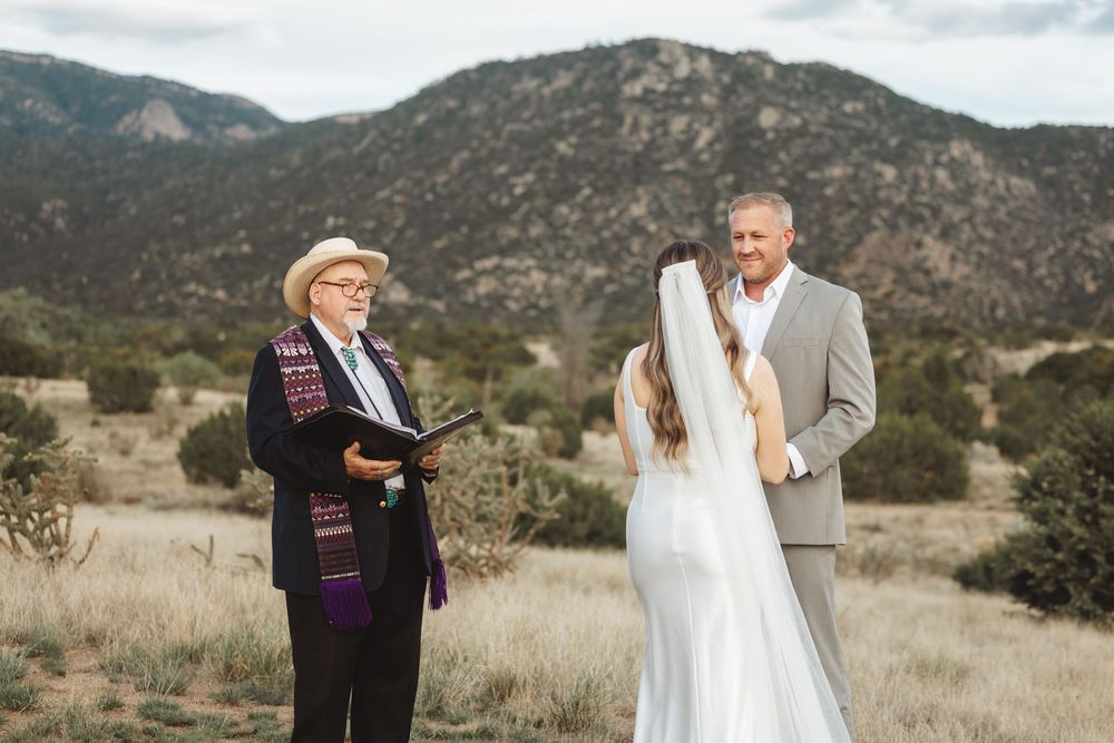 Outdoor wedding ceremony with bride, groom, and officiant against scenic New Mexico mountain backdrop.