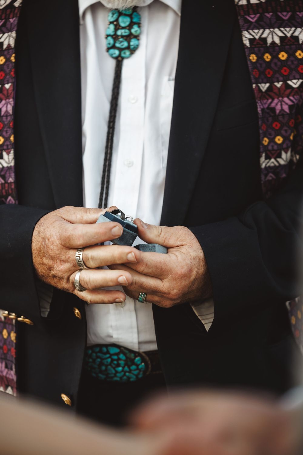 Elderly man in black suit with turquoise bolo tie and rings holding a small object during a wedding ceremony.