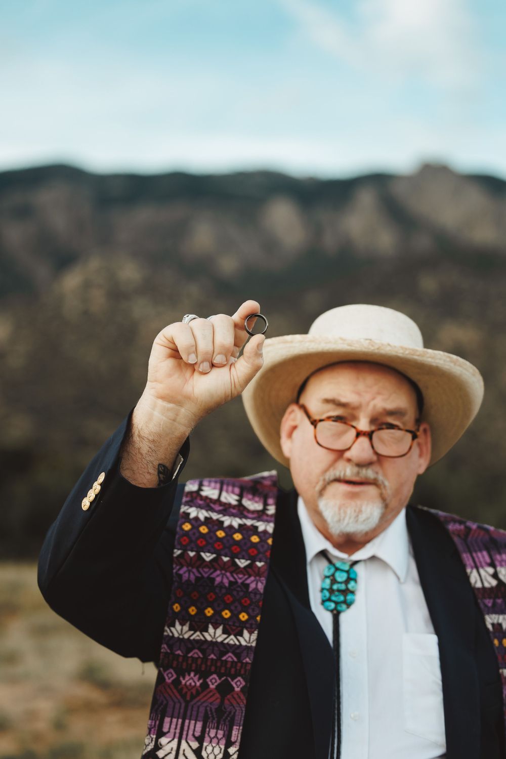 Elderly man in straw hat, glasses, and turquoise bolo tie holds a ring aloft outdoors with mountains behind him.
