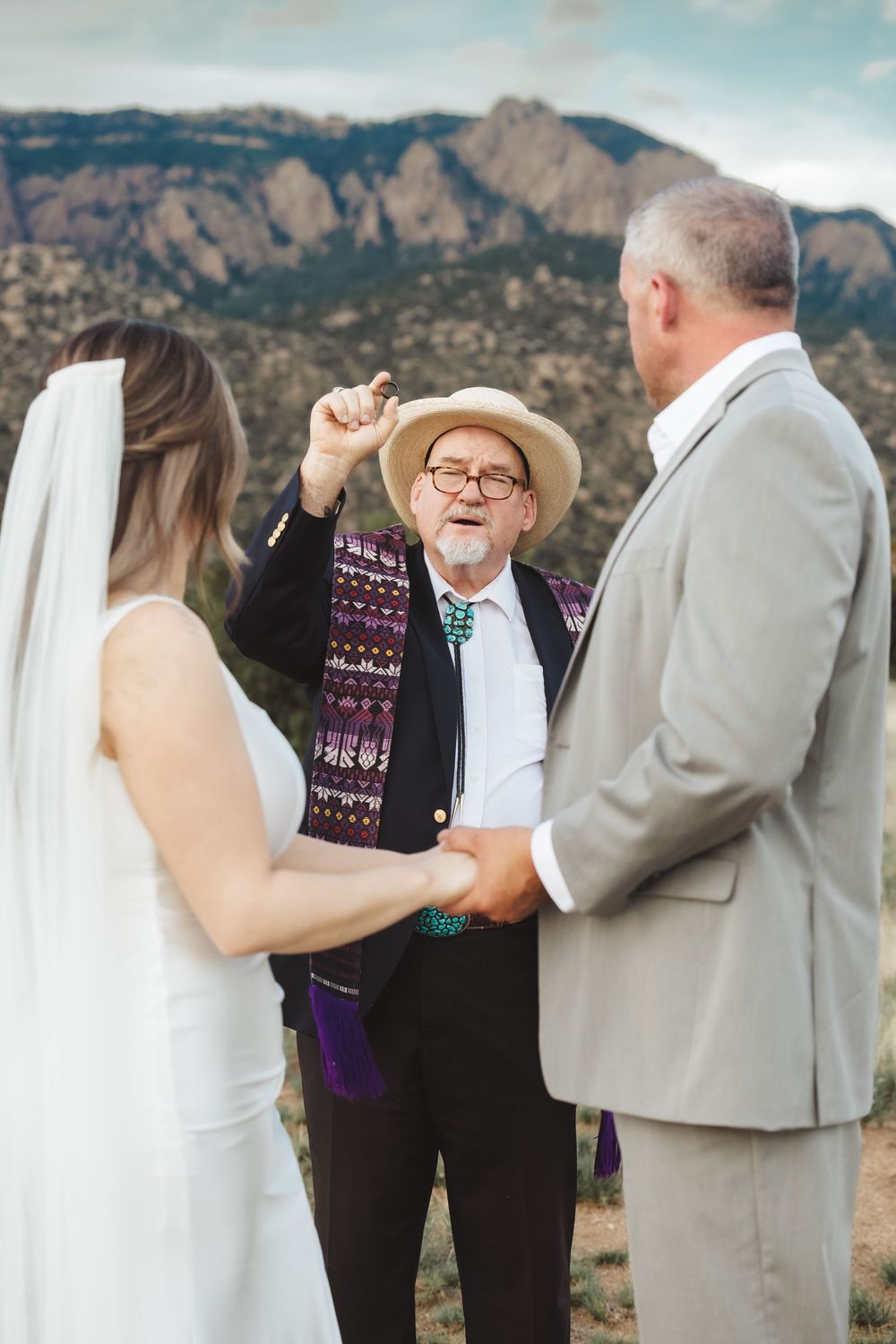 Officiant in straw hat gestures during outdoor mountain wedding ceremony with bride and groom holding hands.