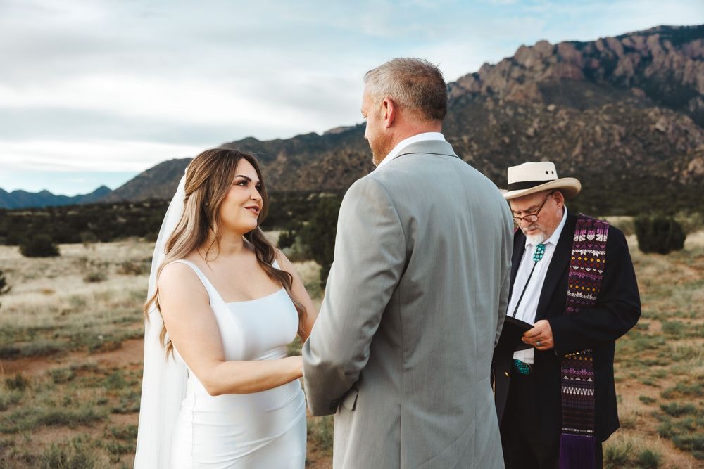 Bride and groom hold hands during outdoor desert wedding ceremony with officiant and mountain backdrop.
