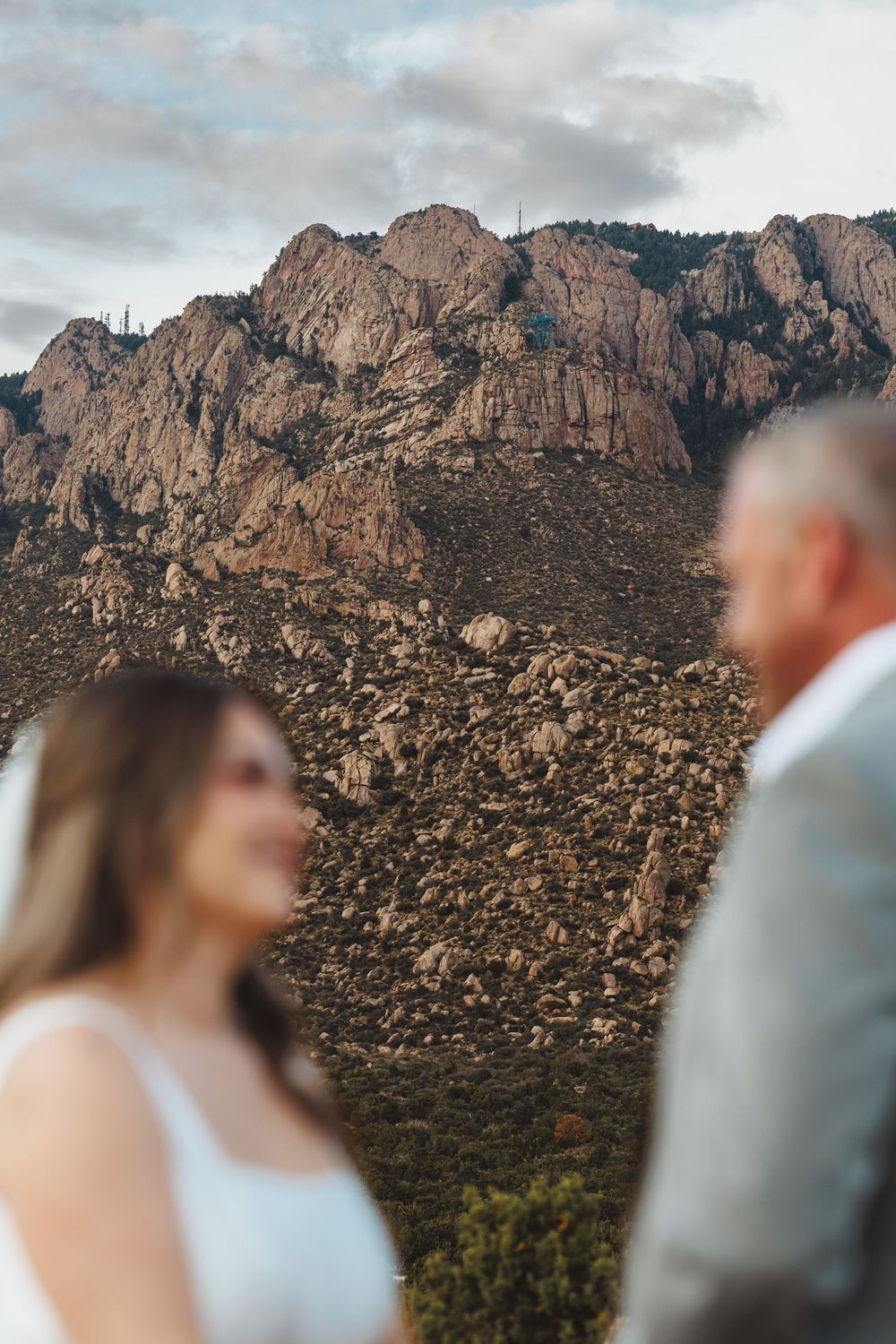 Couple in wedding attire stands before rugged rocky mountain landscape under cloudy sky at golden hour.