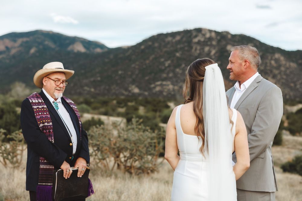 Outdoor wedding ceremony with bride, groom, and officiant against a scenic mountain backdrop in a desert landscape.